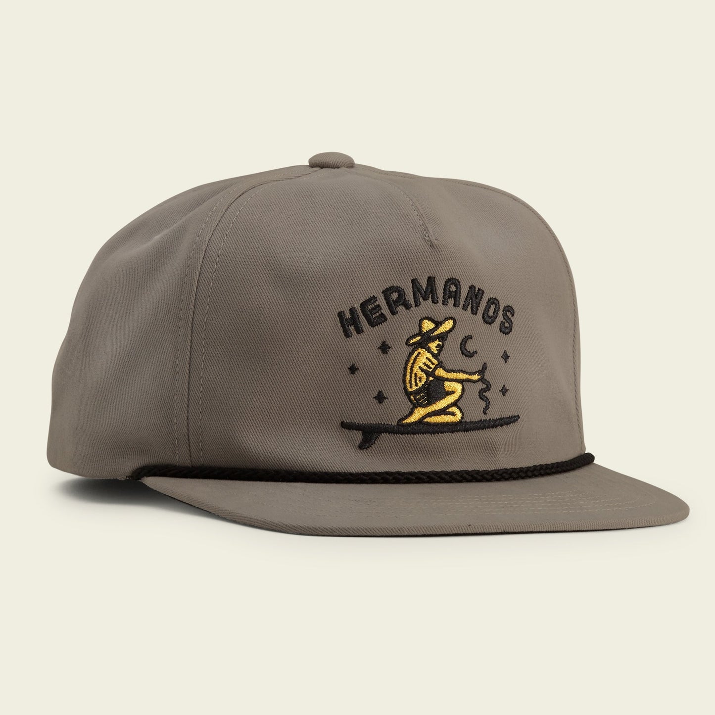 A gray Unstructured Snapbacks hat features an embroidered yellow dog in a cowboy hat sitting on a log with stars, a crescent moon, and “HERMANOS” above. The soft crown and adjustable snapback closure ensure a comfortable fit. - View 1