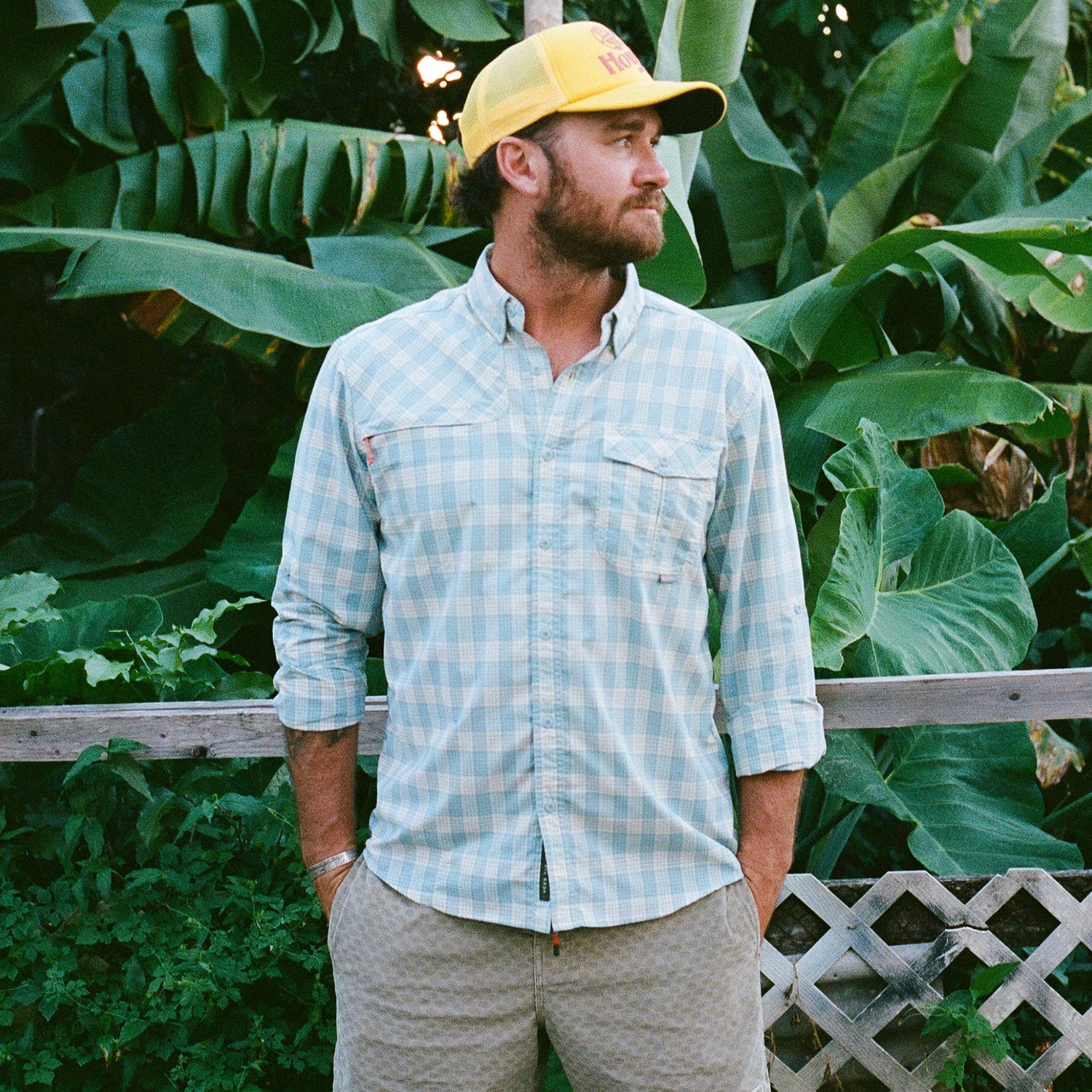 A man with a beard wearing a yellow cap, beige shorts, and a quick drying Matagorda Shirt stands in front of lush green plants and a wooden fence, looking off to the side. - View 2