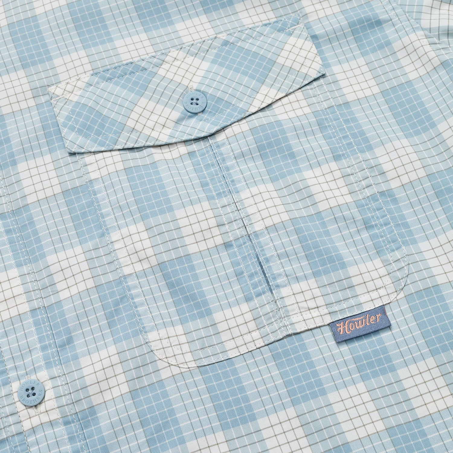 Close-up of a light blue and white checkered Matagorda Shirt with a buttoned chest pocket. The pocket features a blue button and a small fabric tag with the word Howler stitched in red. Ideal as a quick drying travel shirt. - View 3