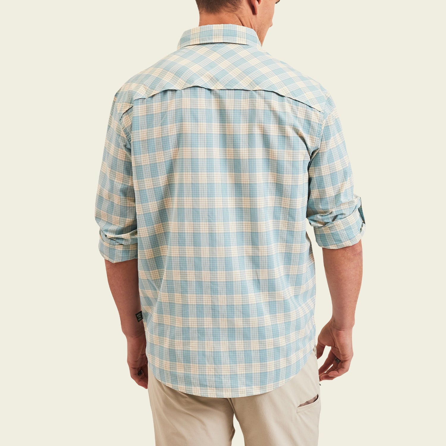 A man is shown from the back wearing a light blue and white plaid Matagorda Shirt with rolled-up sleeves and khaki pants, standing against a plain light background. This travel shirt is quick drying, perfect for any adventure. - View 5