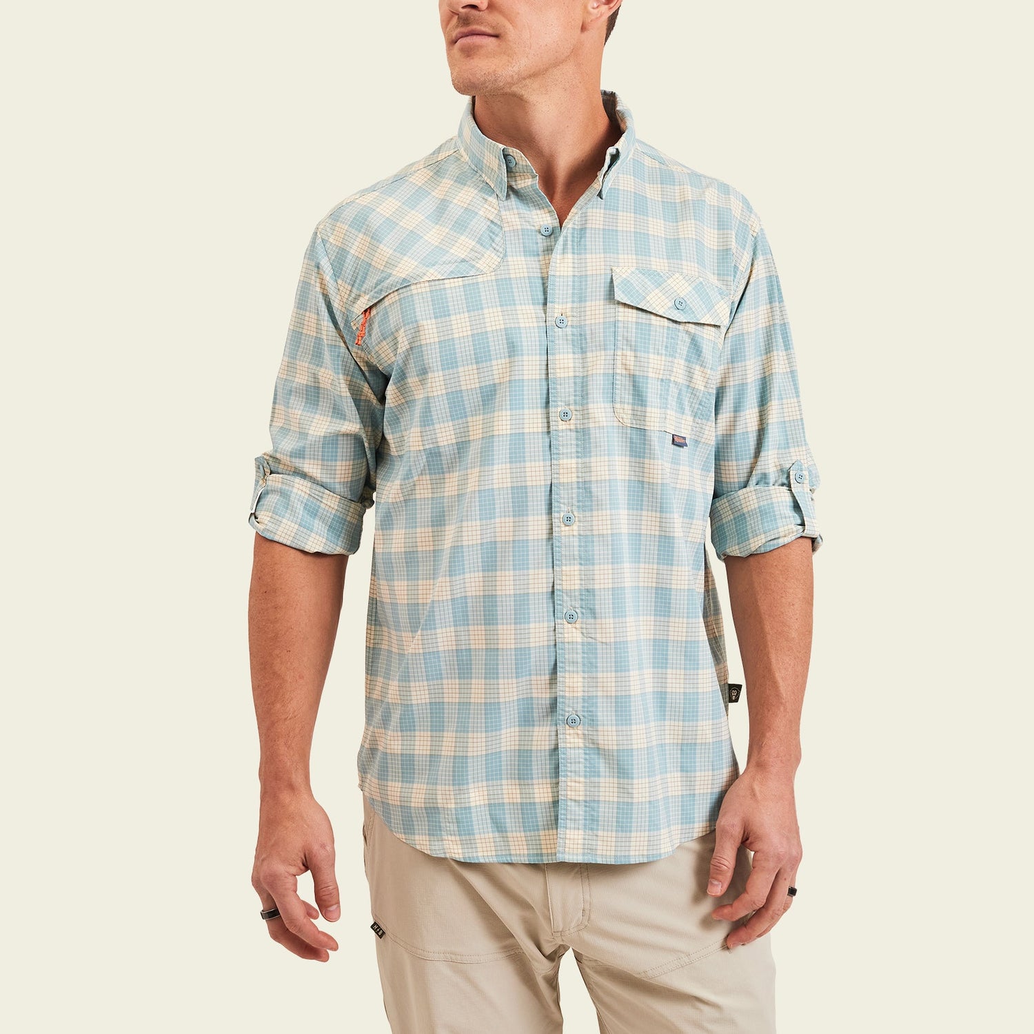 A man wearing a light blue and beige plaid Matagorda Shirt with rolled-up sleeves and khaki pants, standing against a plain off-white background. This stylish travel shirt is quick drying, perfect for adventures on the go. - View 4