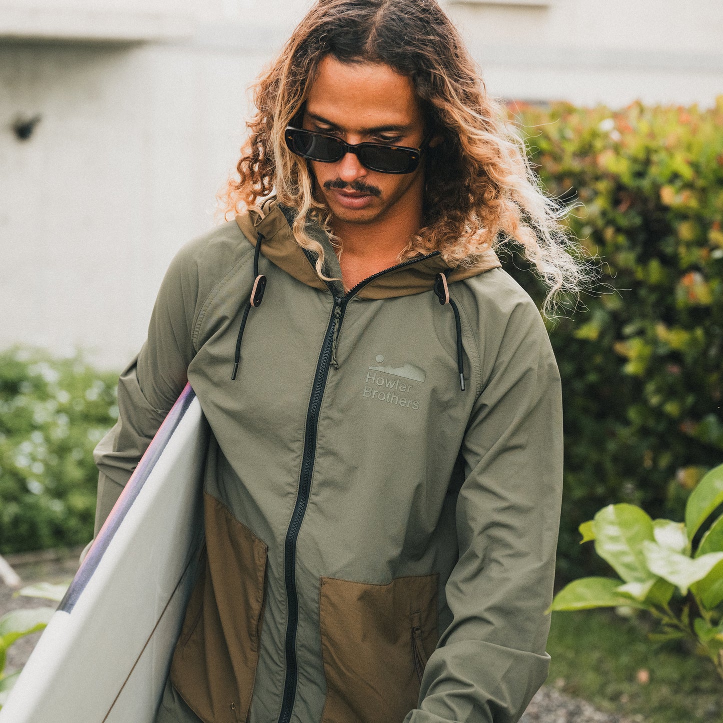 A person with long, wavy hair wearing sunglasses and a green lightweight windbreaker holds a surfboard while standing outdoors near greenery. - View 2