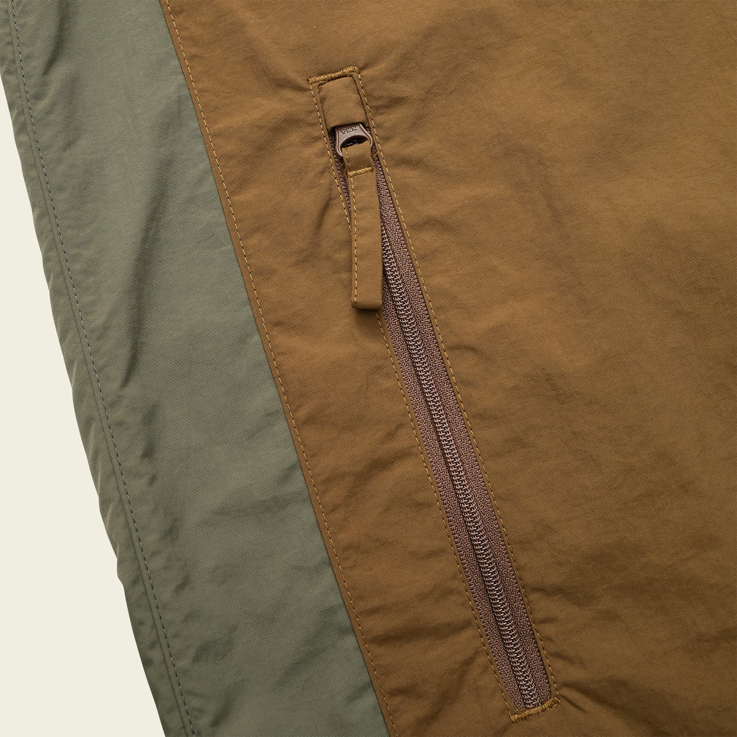 Close-up of a brown zippered pocket on the Seabreacher Jacket, with olive green fabric on the left side. The lightweight windbreaker material appears sturdy and textured, and the zipper pull is clearly visible. - View 6