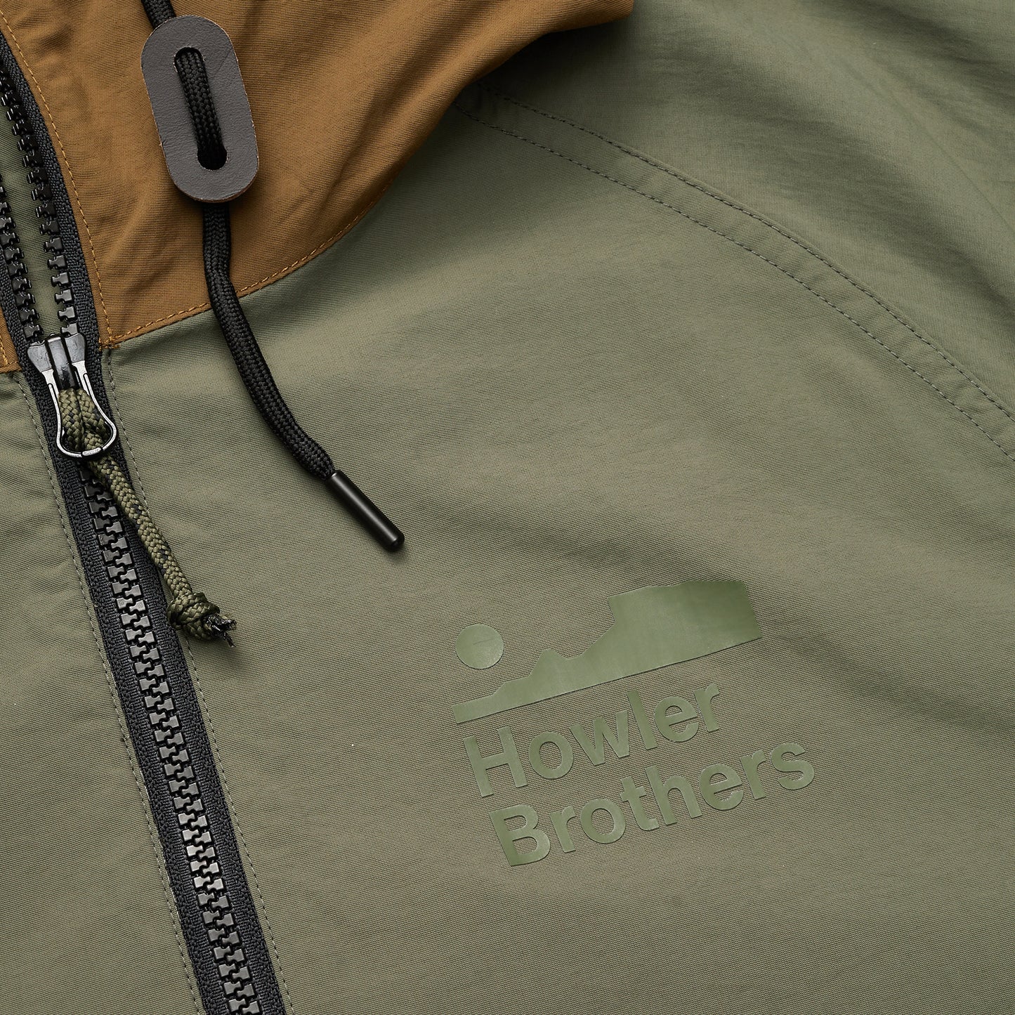 Close-up of the Seabreacher Jacket, a lightweight windbreaker in green and brown with a partially zipped zipper. The Howler Brothers logo is printed near the zipper, alongside a black drawstring with toggle. Water resistant fabric keeps you comfortable. - View 3