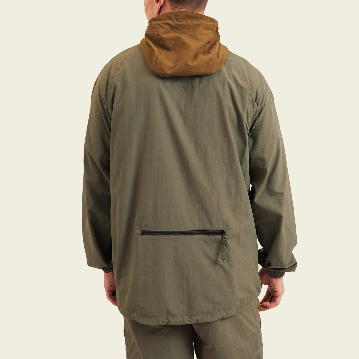 A person wearing an olive green, water resistant Seabreacher Jacket with a brown hood, seen from the back. The jacket features a zipper pocket near the lower back and is paired with matching pants. The background is plain and light-colored. - View 5