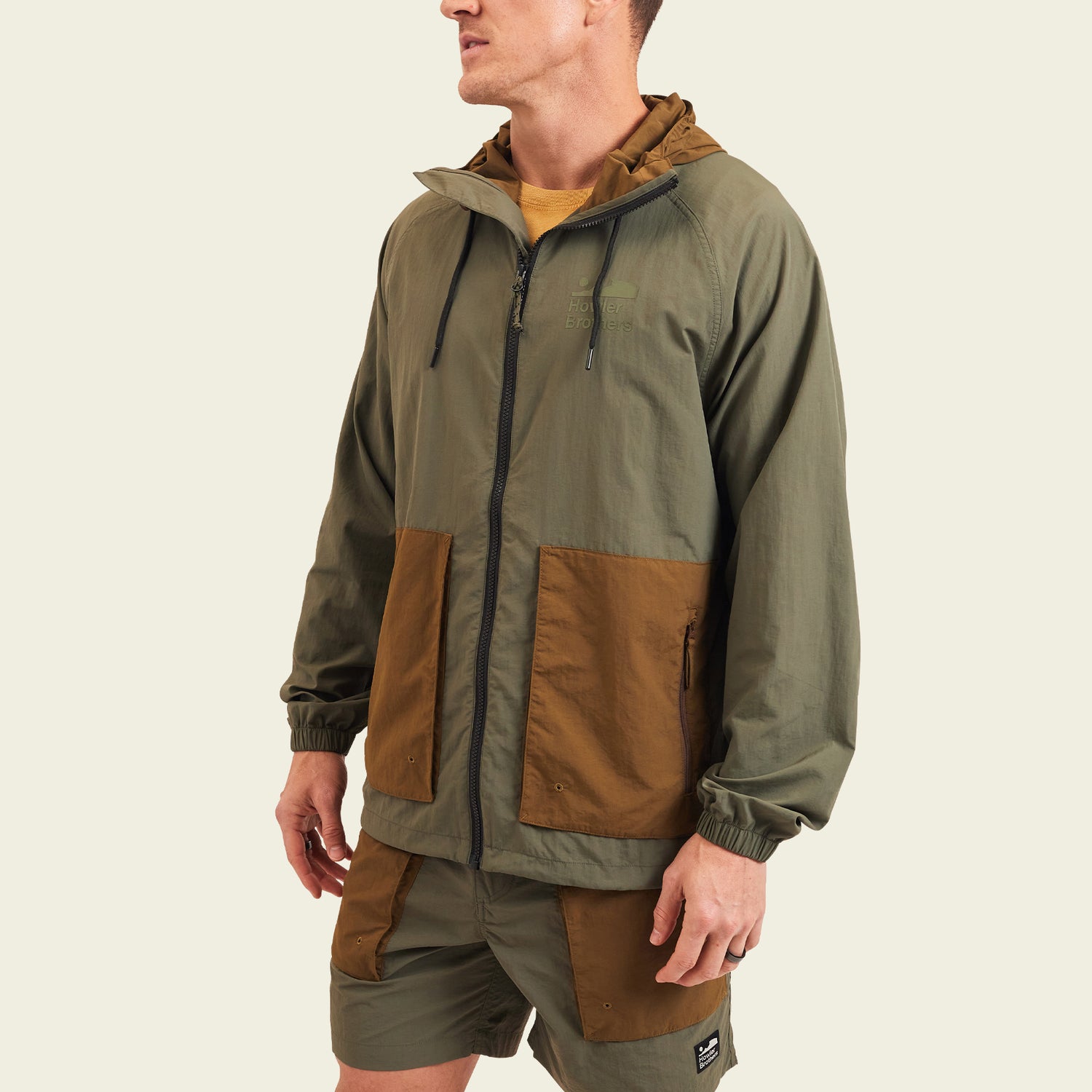 A man wearing a Seabreacher Jacket—an olive green and brown lightweight windbreaker with matching shorts—stands facing slightly left. The water-resistant jacket features a zip front, drawstring hood, and large front pockets. Background is plain and light-colored. - View 4