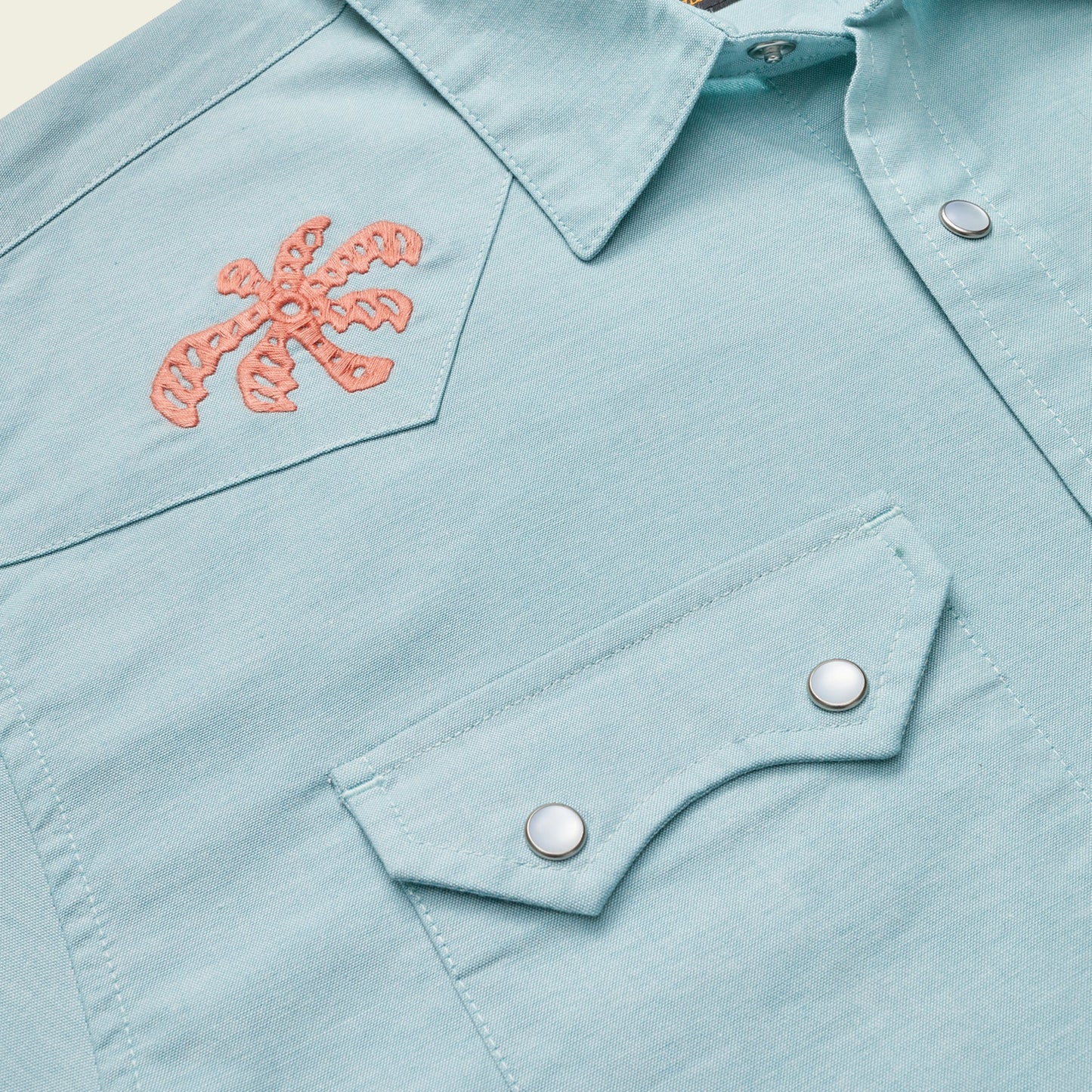 Close-up of a light blue Crosscut Deluxe Shortsleeve summer shirt with pointed collar and snap-button chest pocket, featuring an orange palm tree motif embroidery on the upper left side. - View 3