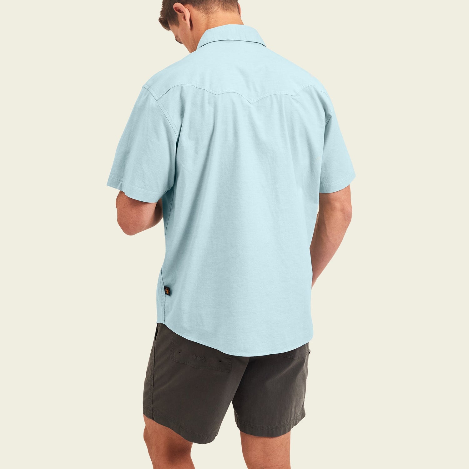 A man stands facing away, wearing a light blue Crosscut Deluxe Shortsleeve summer shirt with dark gray shorts against a plain light background. - View 5
