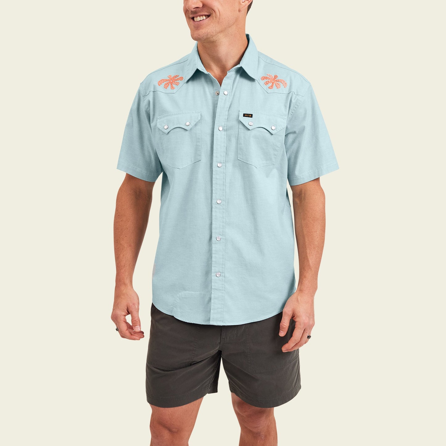 Smiling man wearing a light blue Crosscut Deluxe Shortsleeve summer shirt featuring orange lobster motif embroidery on the shoulders and dark gray shorts, standing against an off-white background. - View 4