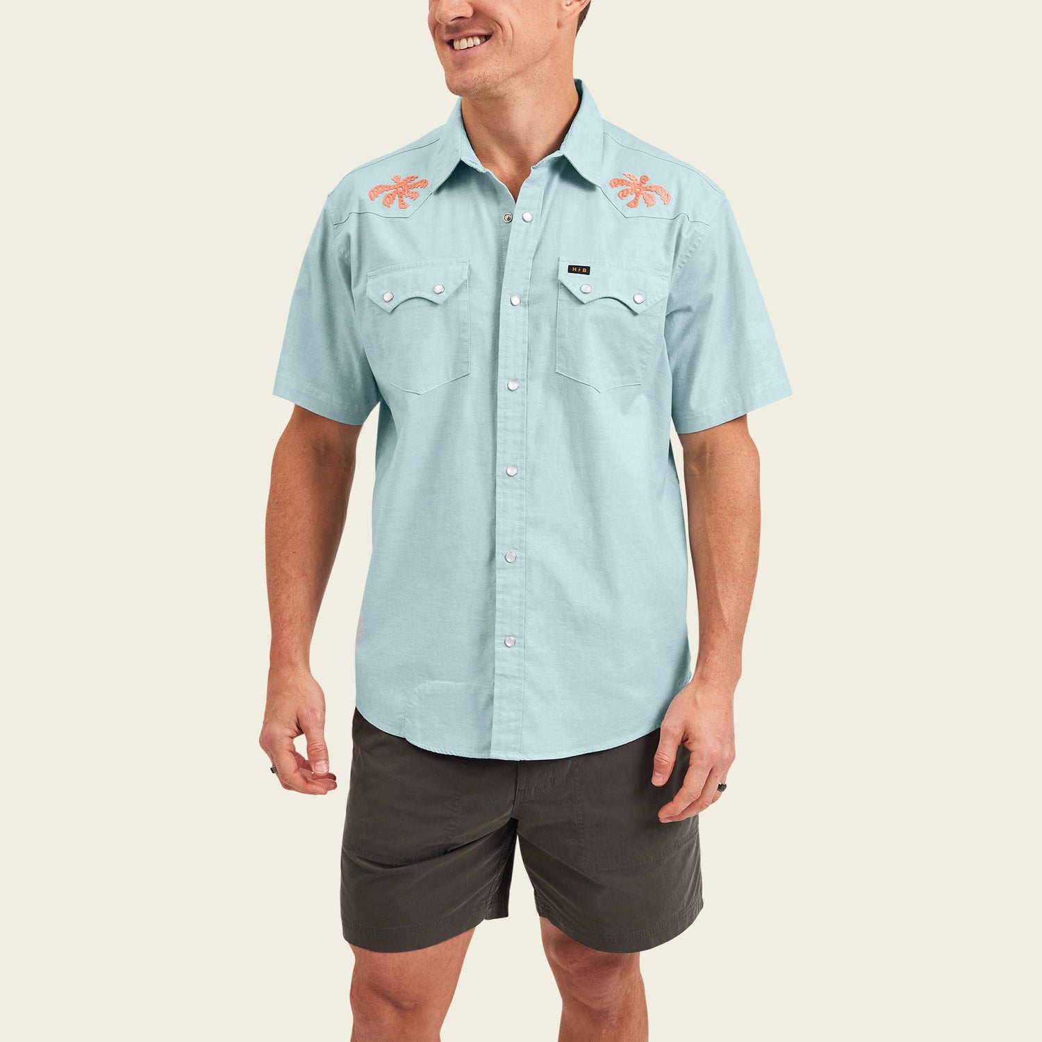 Smiling man wearing a light blue Crosscut Deluxe Shortsleeve summer shirt featuring orange lobster motif embroidery on the shoulders and dark gray shorts, standing against an off-white background. - View 4