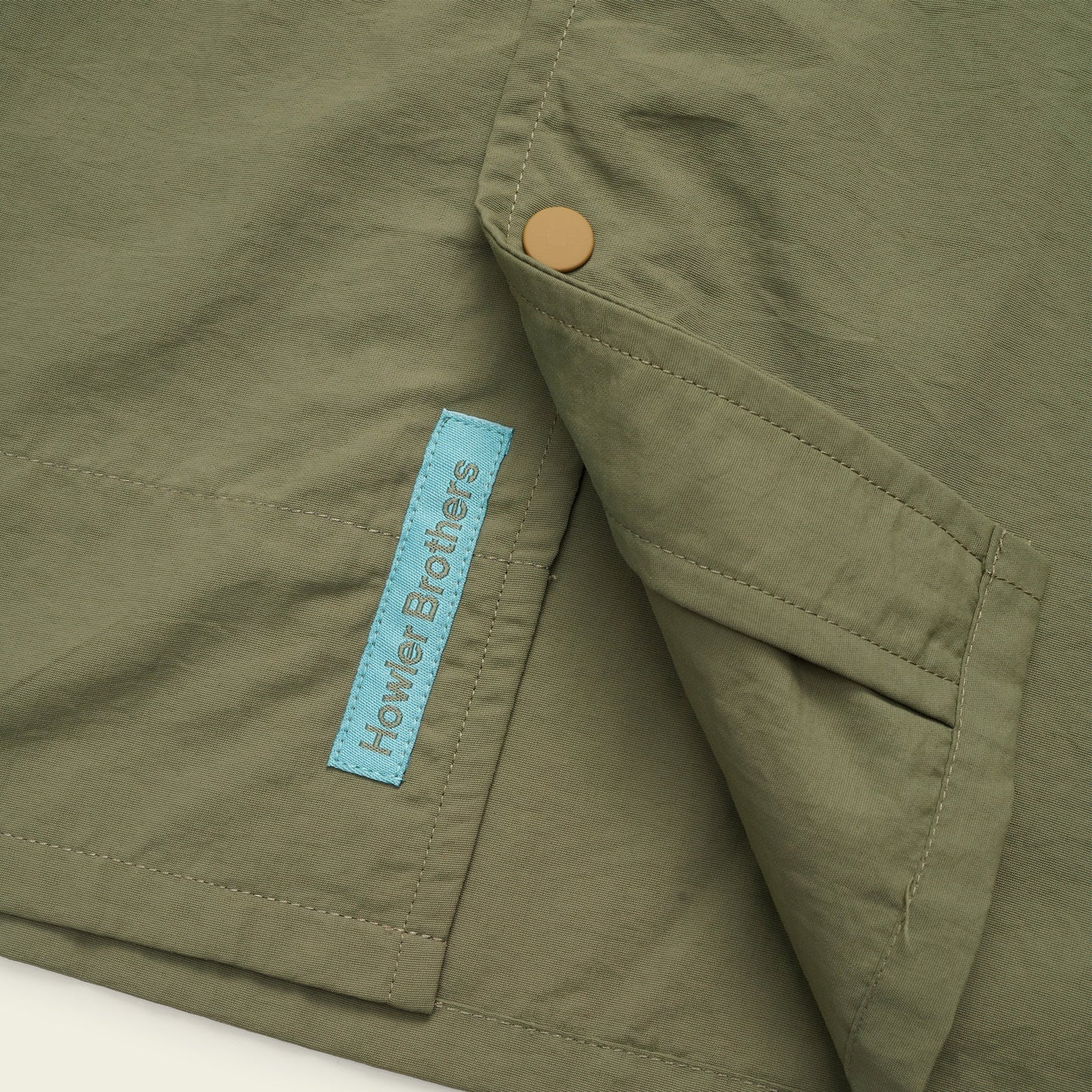 Close-up of an olive green fabric with a brass snap button and a blue Howler Brothers tag, likely part of the Forager Utility Shirt—perfect for hiking trails, featuring visible seams and a pocket for added utility. - View 7