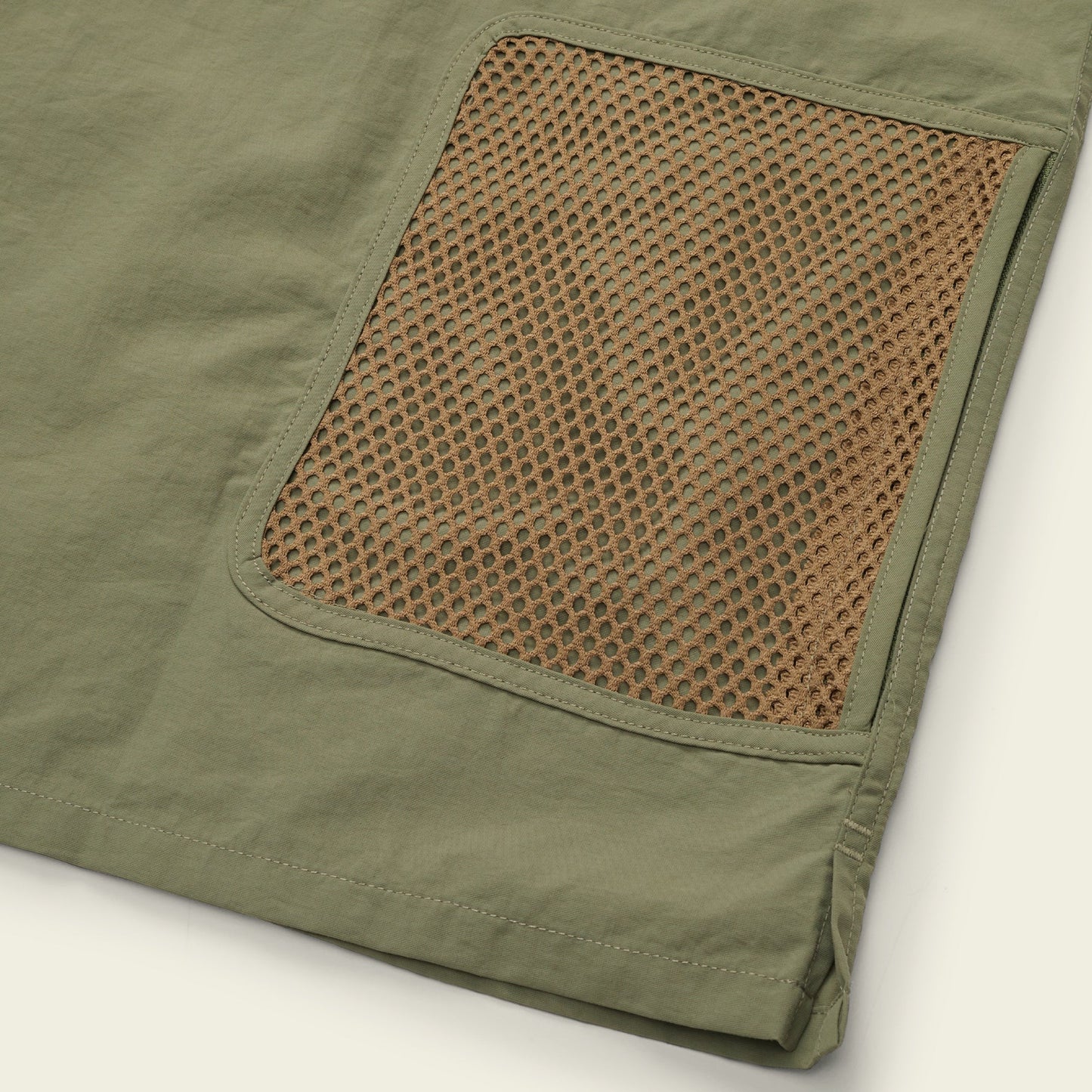 Close-up of an olive green Forager Utility Shirt fabric with a brown mesh pocket sewn on top, showing the texture and stitching details—perfect for comfort and storage on hiking trails. The edge of the fabric is visible in the bottom right corner. - View 3
