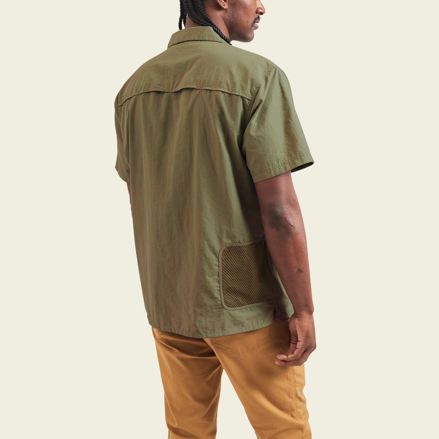 A man stands facing away, wearing the olive-green Forager Utility Shirt with mesh pocket detail—an ideal tech shirt for hiking trails—paired with tan pants, against a plain light background. - View 5