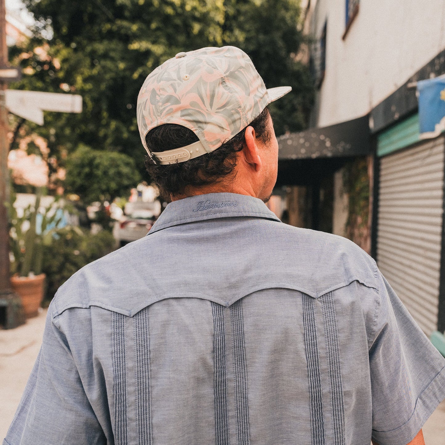 A man in a light blue Guayabera with unique embroidery and a patterned cap walks down a sidewalk, facing away from the camera. Trees, buildings, and a closed storefront appear in the background. - View 2
