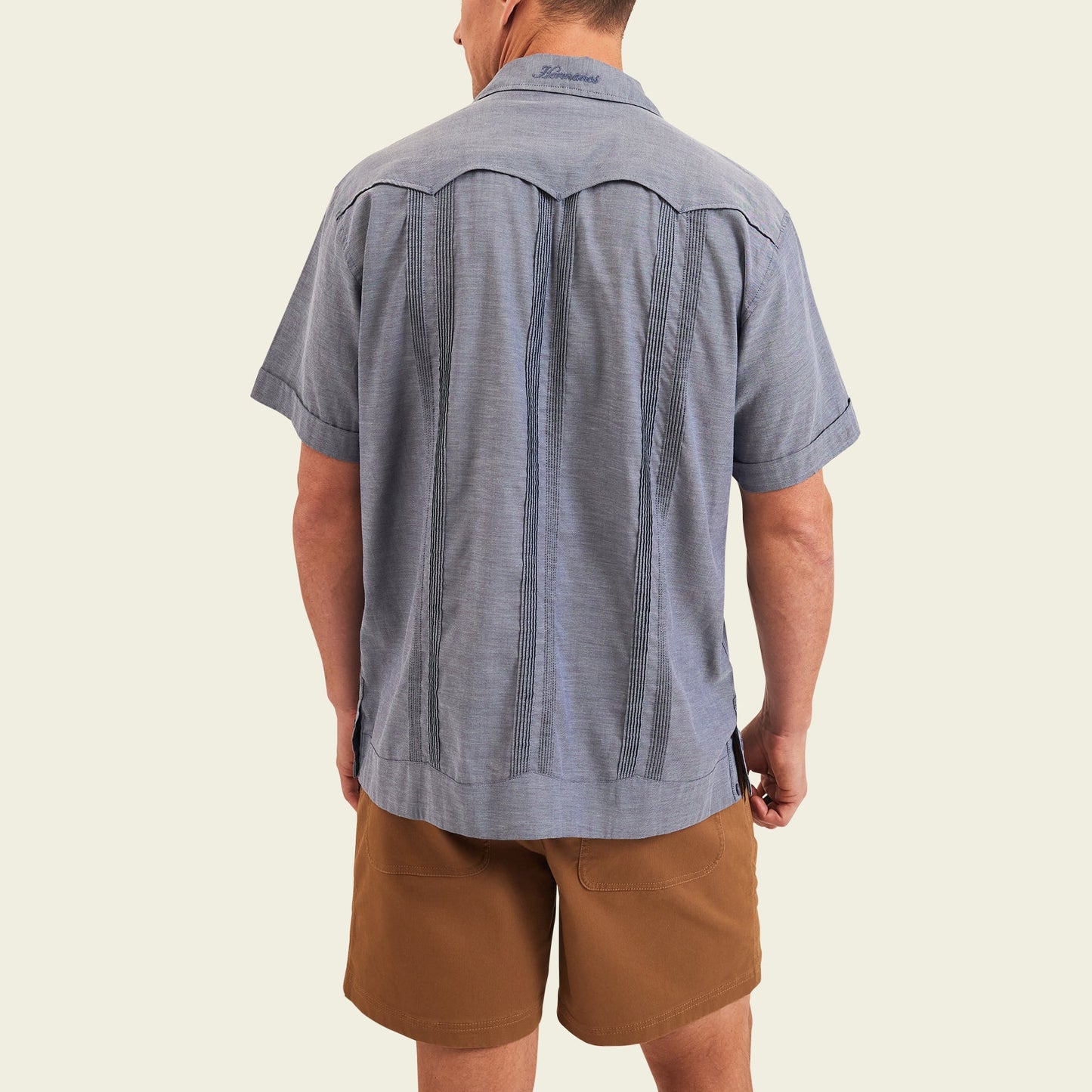 A man stands facing away from the camera, wearing a short-sleeve blue patterned Guayabera shirt with unique embroidery and brown shorts, set against a plain light background. - View 5