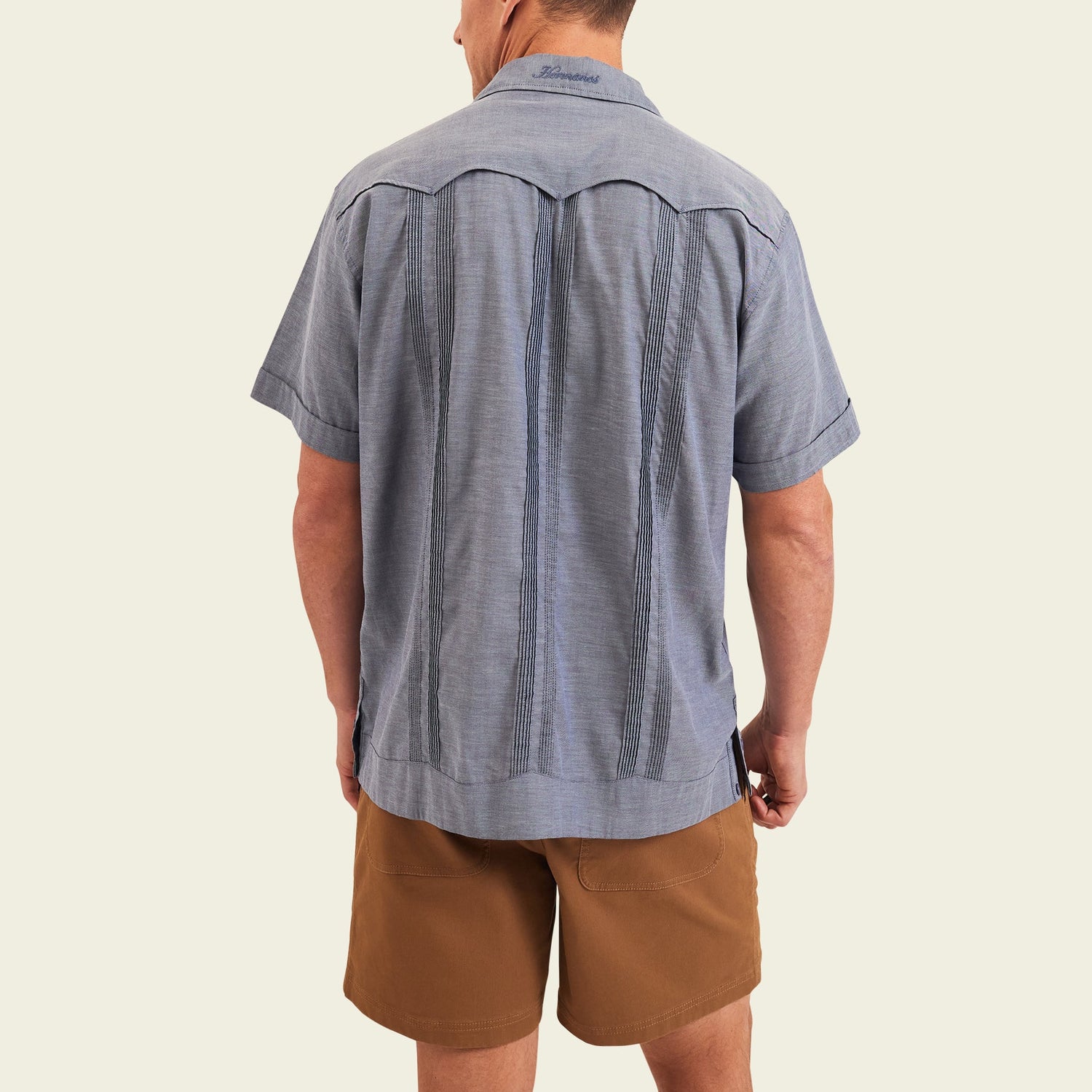 A man stands facing away from the camera, wearing a short-sleeve blue patterned Guayabera shirt with unique embroidery and brown shorts, set against a plain light background. - View 5