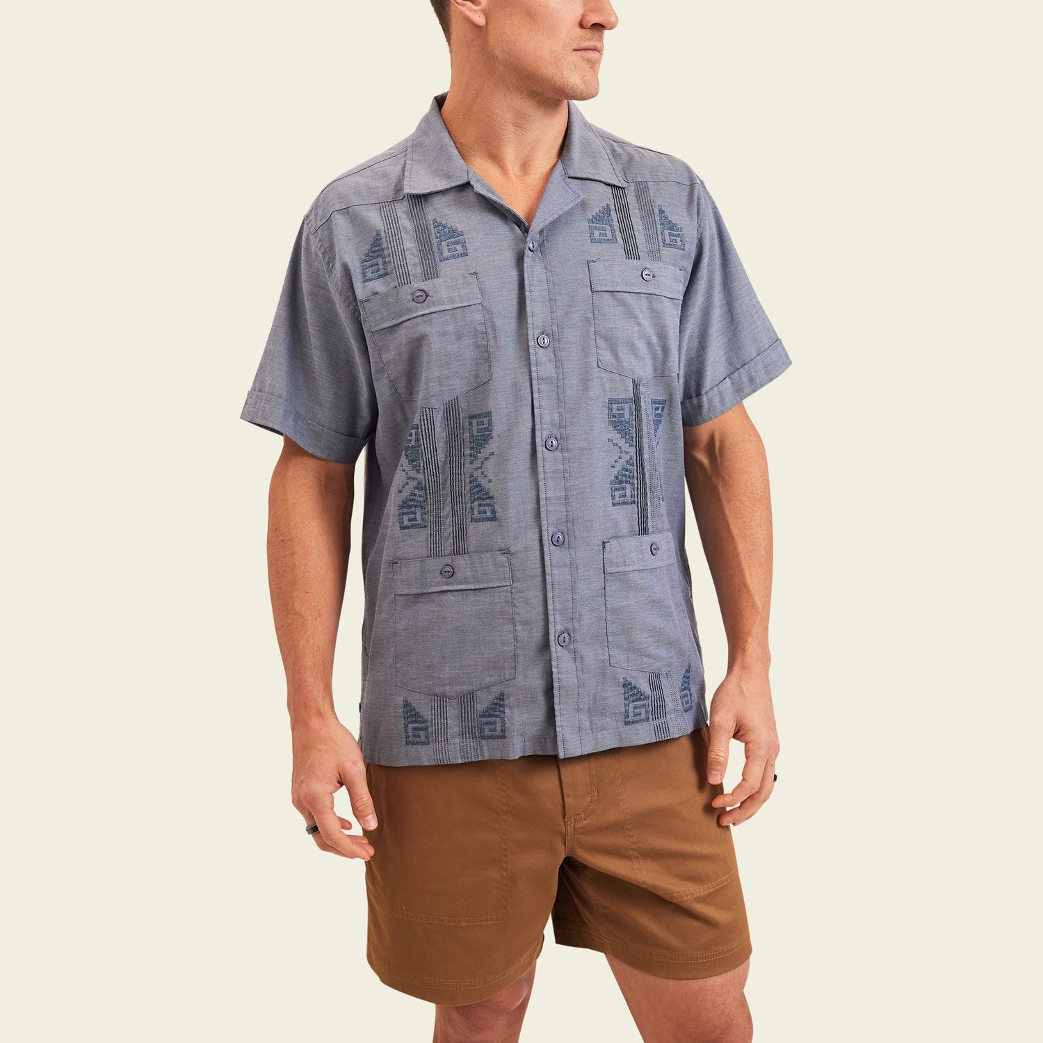 A person wearing a short-sleeve, button-up blue patterned Guayabera with unique embroidery and brown shorts stands against a plain, light background, looking off to the side. - View 4