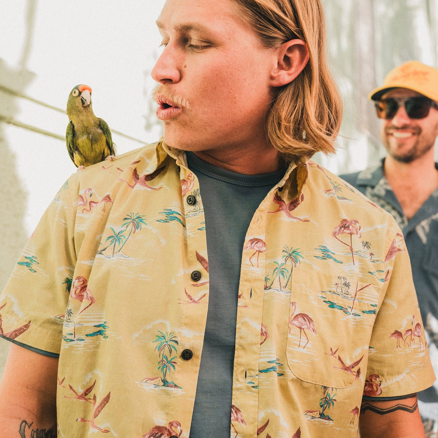 A man with long blond hair wearing a Mansfield Shirt in unique seasonal patterns looks at a parrot perched on his shoulder. Another man in sunglasses and a yellow cap smiles in the background.