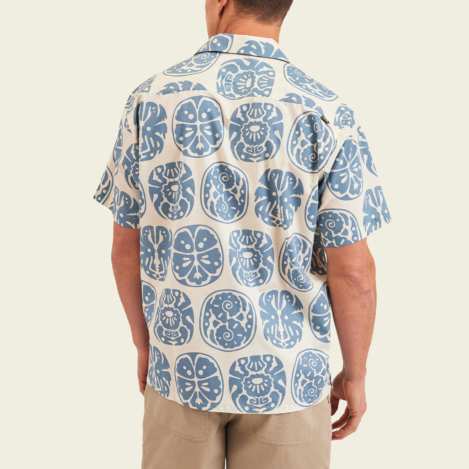 A man wears a short-sleeve, camp collar shirt with a blue and white circular Sea Idols print, reminiscent of vintage Hawaiian shirts; he stands facing away in light khaki shorts, highlighting the shirt’s back. - View 5