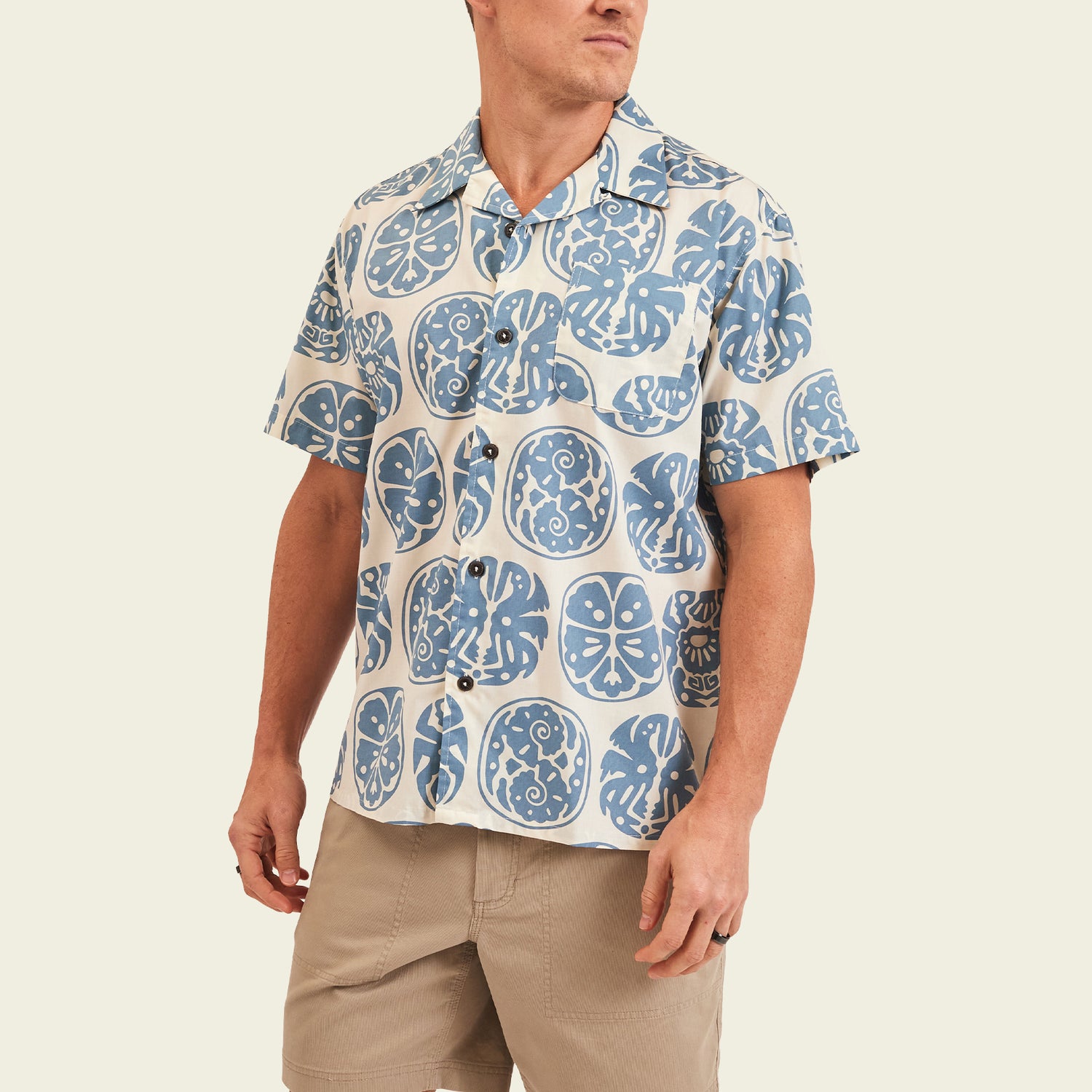 A man wears a short-sleeve, camp collar shirt featuring a blue and white tropical Sea Idols print with floral motifs, paired with light tan shorts, standing against a plain off-white background. - View 4