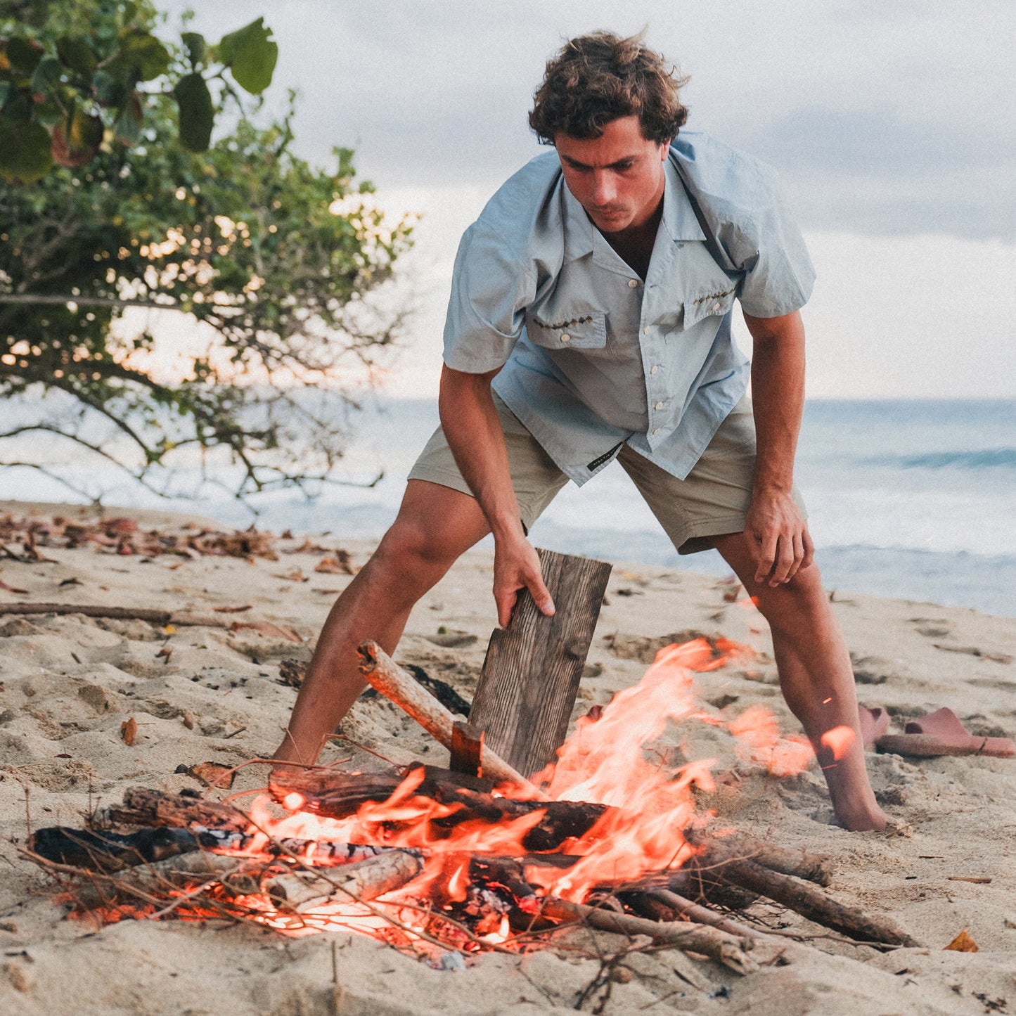 A man in a light blue shirt and cotton twill shorts adds a piece of wood to a campfire on a sandy beach, with the ocean and trees visible in the background. - View 2