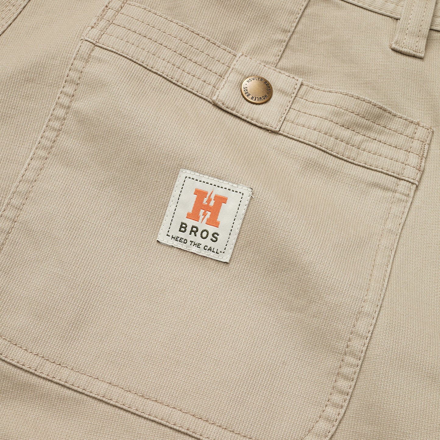 Close-up of beige cotton twill shorts featuring a front pocket with a metal button and a white label that reads “H BROS HEED THE CALL” in black text, with an orange “H” logo above it. - View 3