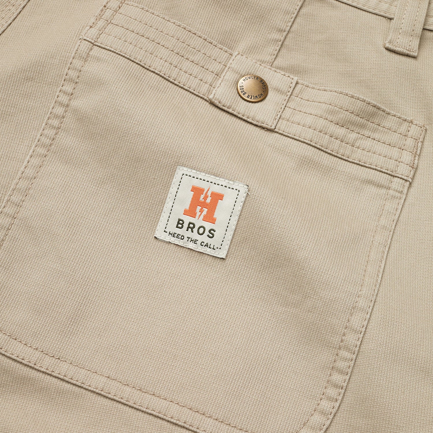 Close-up of beige cotton twill shorts featuring a front pocket with a metal button and a white label that reads “H BROS HEED THE CALL” in black text, with an orange “H” logo above it. - View 3