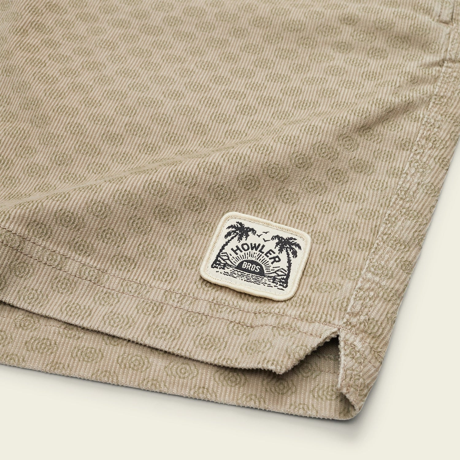 Close-up of tan Pressure Drop Cord Shorts made from stretch cotton corduroy, featuring an elastic waistband and a stitched Howler Bros label with a palm tree graphic near the hem. - View 3