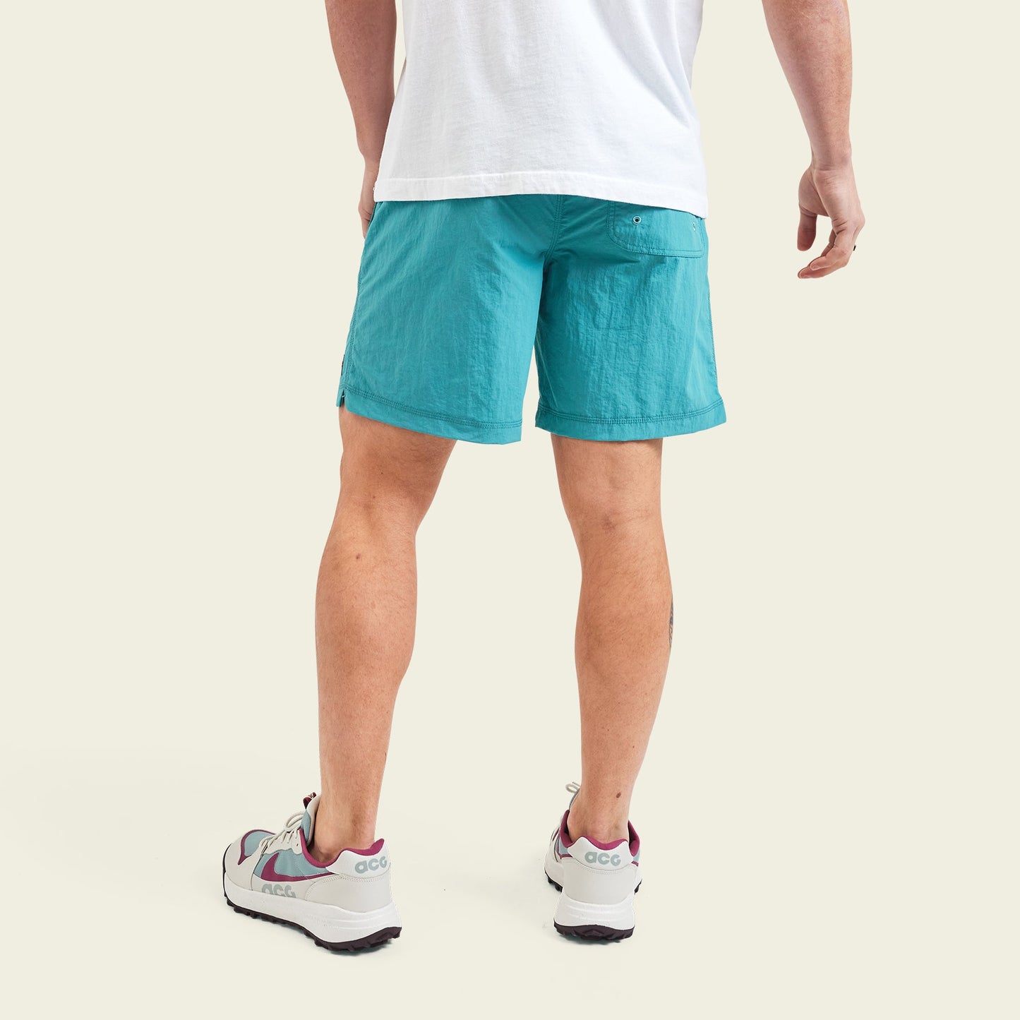 A person stands facing away, wearing a white T-shirt, quick drying nylon running and hiking shorts in teal, and white sneakers with maroon accents, set against a light background. - View 4