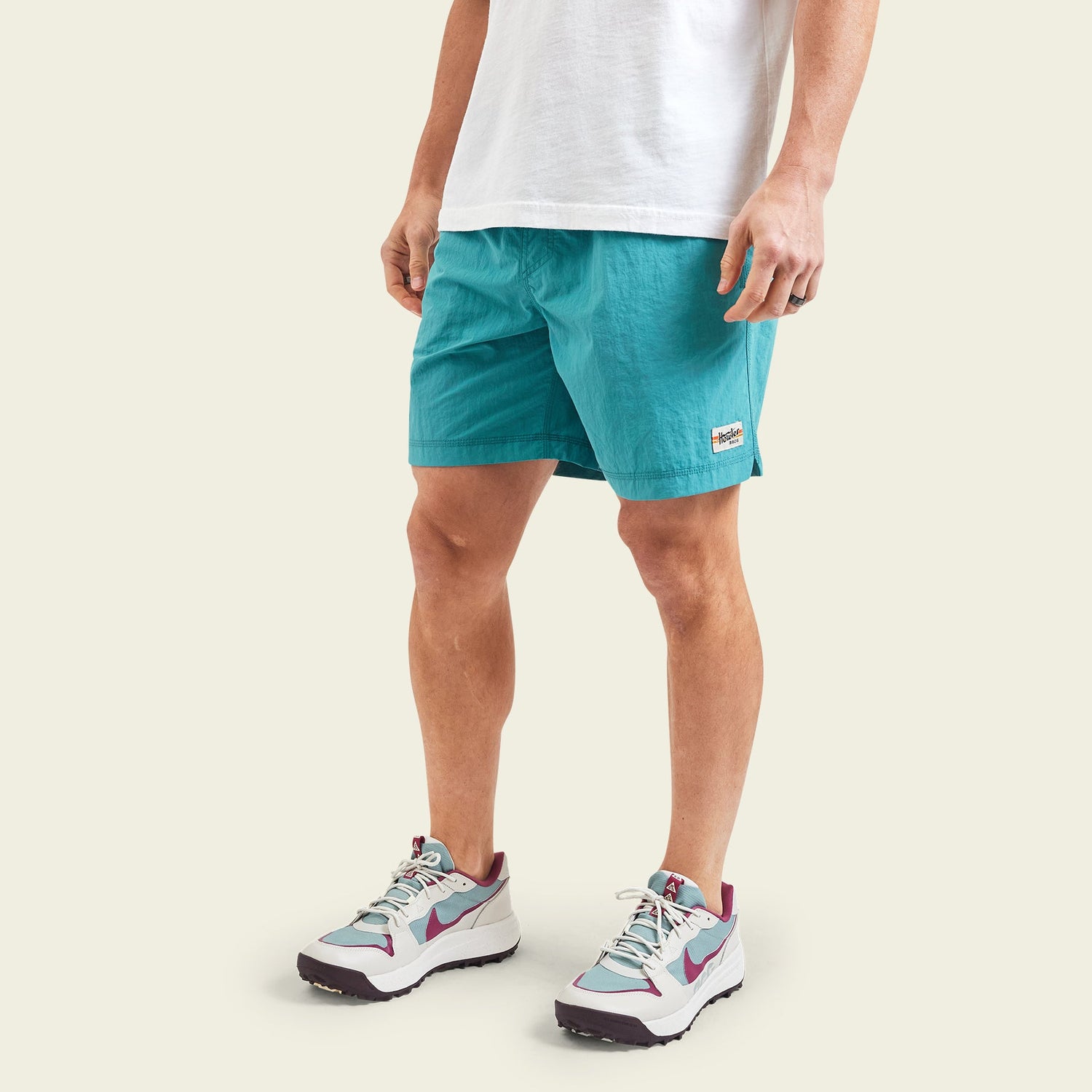 A person wearing a white t-shirt, running and hiking shorts in teal with an elastic waistband, and white sneakers with maroon and gray details, stands against a plain light background. - View 3