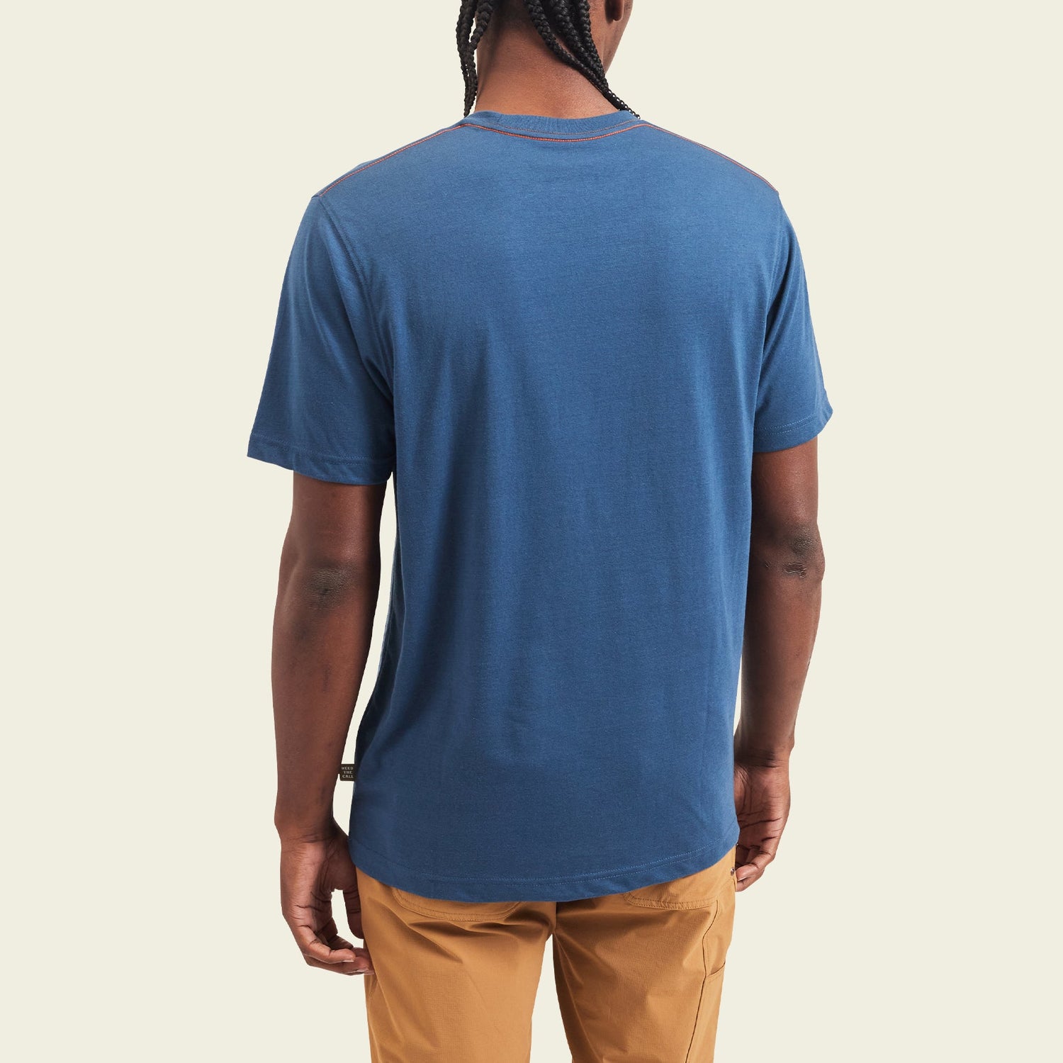 A person with braided hair is shown from behind, wearing a blue short-sleeve Howler blended t-shirt and tan pants, standing against a plain light-colored background. - View 4