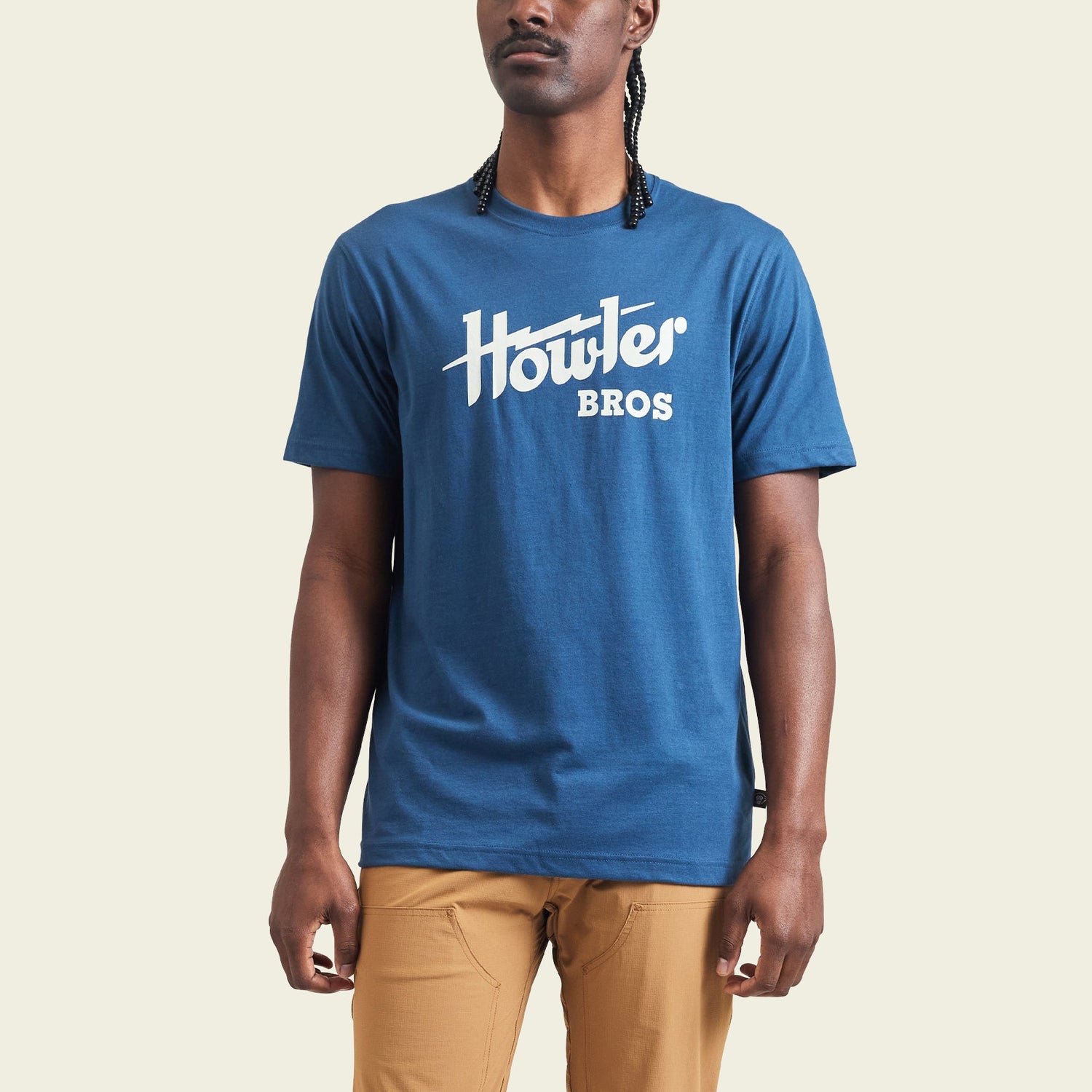 A person wearing a blue cotton-poly blend Howler Bros t-shirt and tan pants stands against a plain, light-colored background. The shirt features a white Howler Bros logo across the chest, showcasing classic Howler designs. - View 3