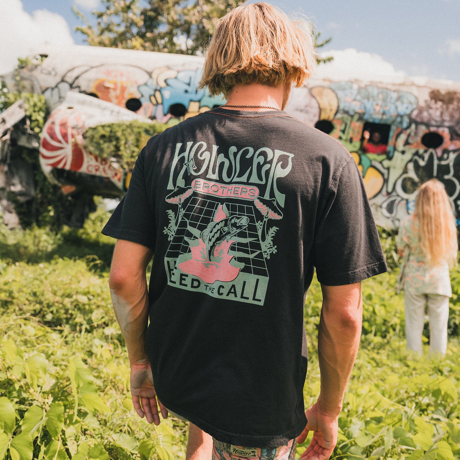 A person with blond hair wearing a black band tee stands in a grassy area near a graffiti-covered abandoned airplane. Another person with long hair is visible in the background, adding to the scene’s experimental artwork vibe.