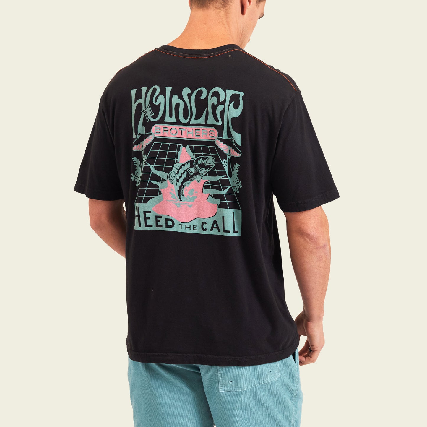 A person wearing a black T-shirt with colorful graphic text reading Howler Brothers Heed the Call and an illustrated fish leaping from water on the back. The design gives off the vibe of vintage t-shirts with experimental artwork. The person wears light blue shorts. - View 4