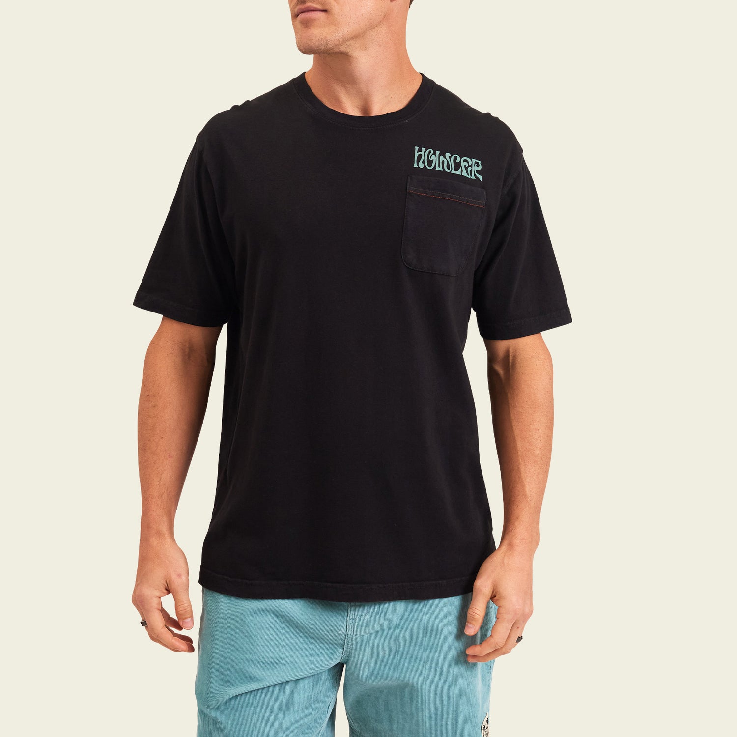 A man wearing a loose-fitting black vintage t-shirt with a chest pocket and blue lettering, paired with light blue shorts, stands against a plain light background. - View 3