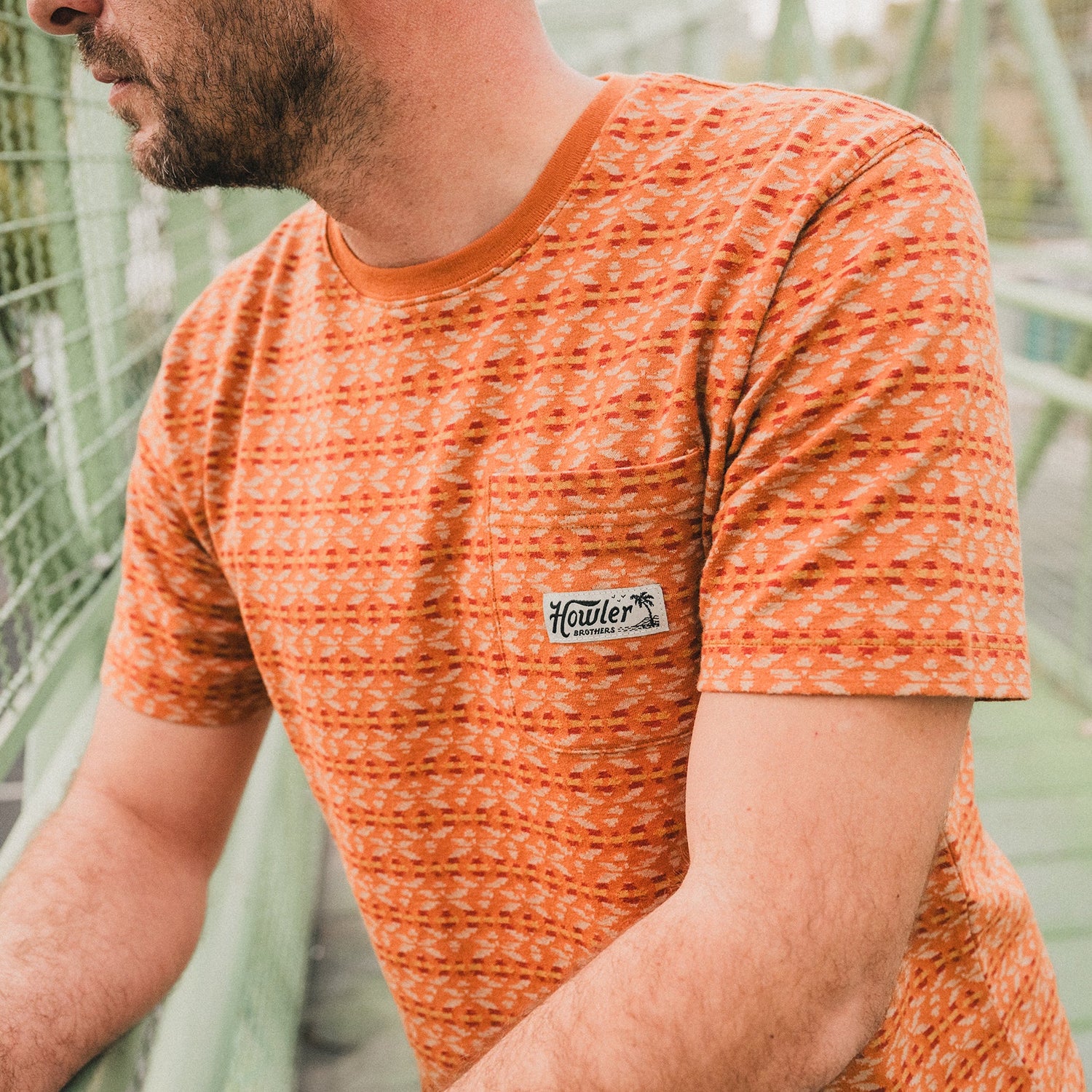 A man wearing an orange patterned cotton jacquard t-shirt with a small chest pocket and a Howler logo stands by a wire fence outdoors, leaning on the railing with his arms. The background is blurred.