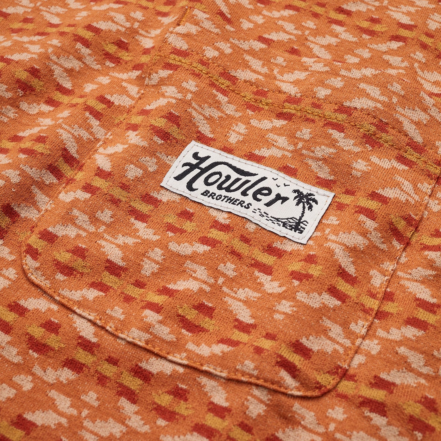 Close-up of an orange patterned cotton jacquard t-shirt with a chest pocket, featuring a white rectangular Howler Brothers label and palm tree graphic. The abstract design in beige and reddish tones evokes a vintage surf shirt vibe. - View 3
