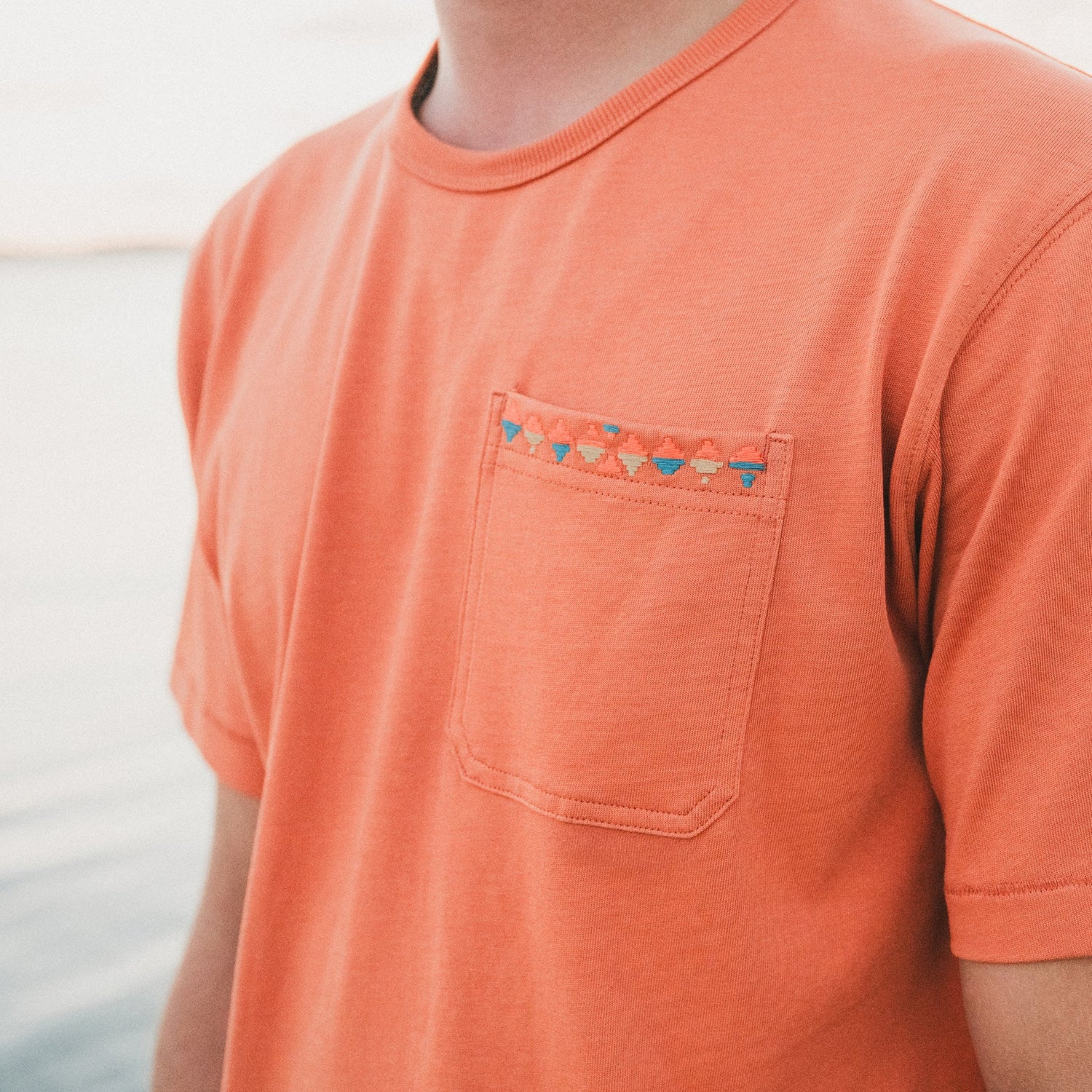 A person wearing a light orange 80s 90s inspired pocket t-shirt with a colorful triangular pattern, standing outdoors near water. The image is cropped to show only the torso and part of the neck.