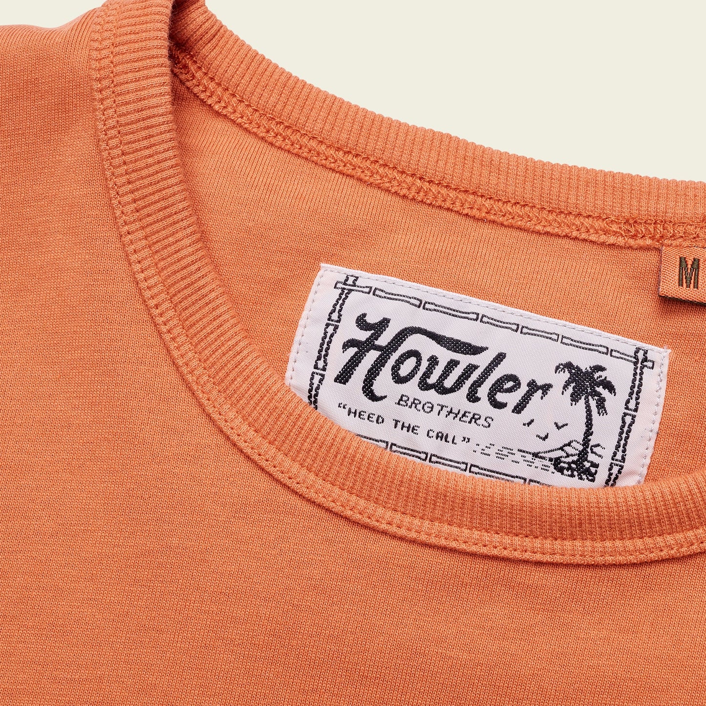 Close-up of an orange heavyweight cotton t-shirt collar with a visible white Howler Brothers label featuring a palm tree illustration and the slogan “Heed the Call.” The shirt’s size “M” tag is partially visible. 80s 90s inspired style. - View 6
