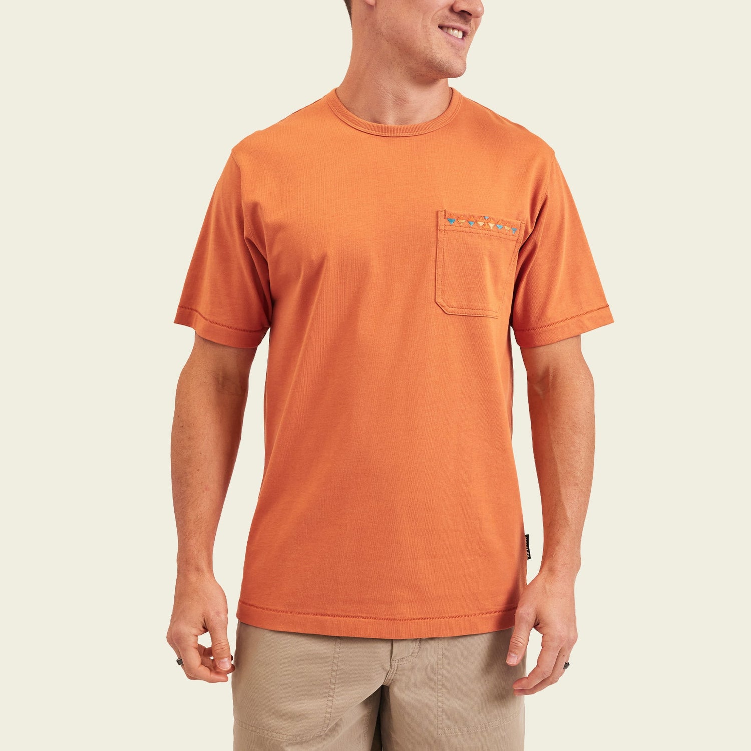 A man wearing a short-sleeve orange heavyweight cotton pocket t-shirt and beige shorts stands smiling against a plain light background. - View 4
