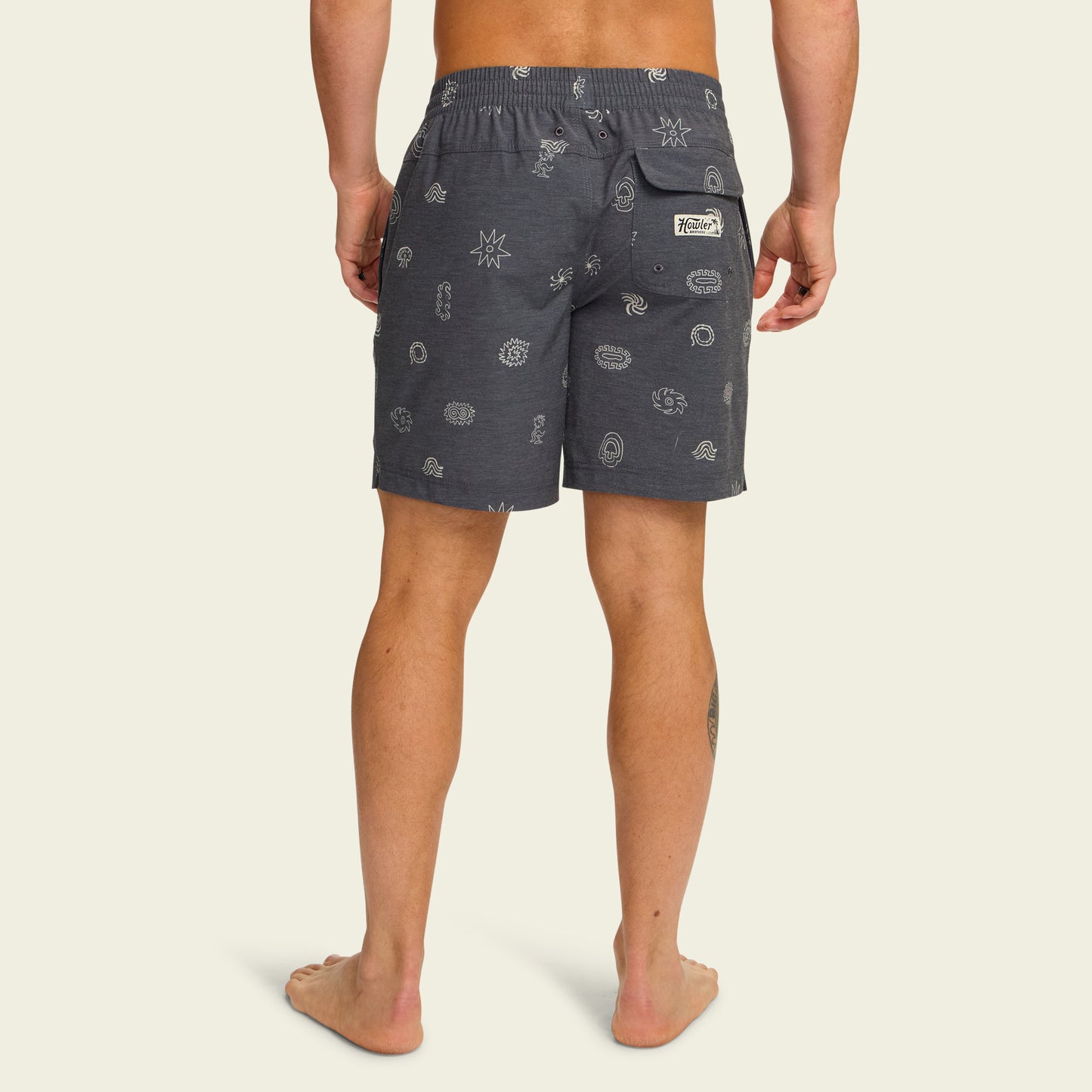 A man stands barefoot facing away, wearing dark gray boardshorts with a subtle pattern, a 7” inseam, an elastic waistband, and a back pocket with a white label, against a plain light background. - View 4