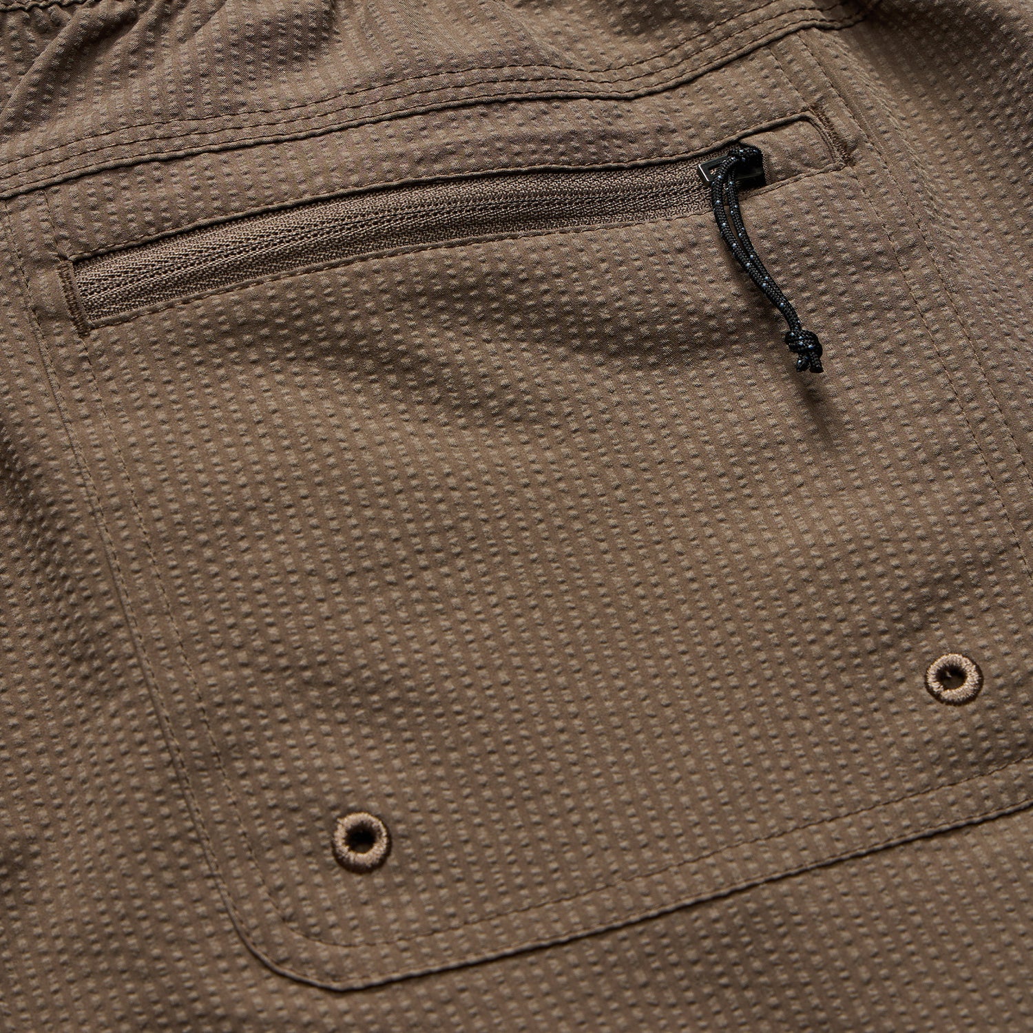 Close-up of tech seersucker shorts in brown textured fabric with a zippered pocket, metal grommets, and visible stitching. The grid-like pattern and drawstring near the zipper highlight their functional design. - View 5