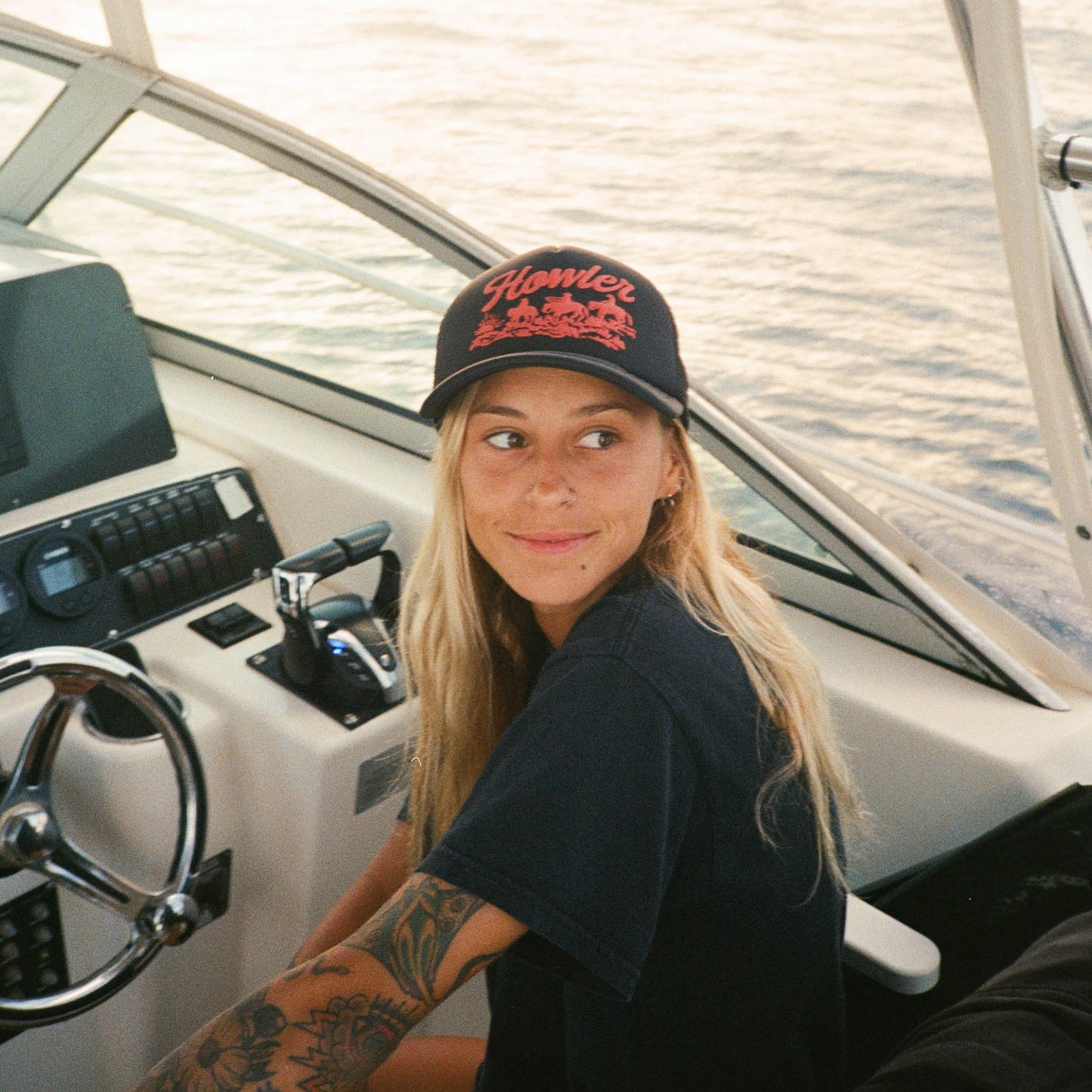 A woman with long blonde hair and tattoos, wearing a black T-shirt and a vintage-inspired high crown hat with a foam front, sits at the helm of a boat, smiling and looking to the side. The background shows calm water. - View 5