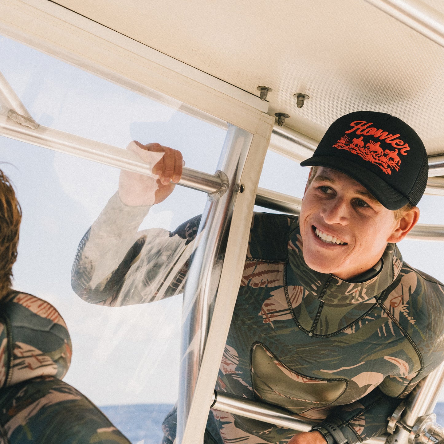 A smiling person in camouflage clothing and a black vintage-inspired hat holds onto a metal bar inside a boat, with sunlight streaming in and another person partially visible beside them. - View 2