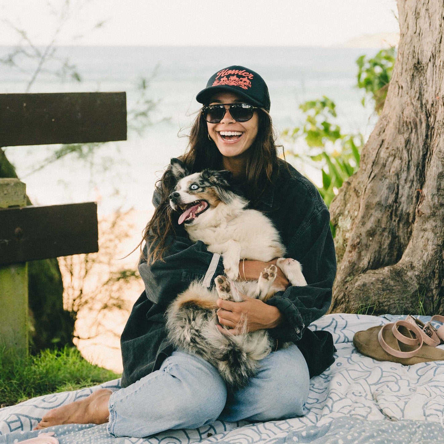A woman wearing sunglasses and a vintage-inspired hat sits on a blanket outdoors, smiling and holding a happy dog in her lap. There are trees, greenery, and water in the background, and sandals rest on the blanket nearby. - View 4