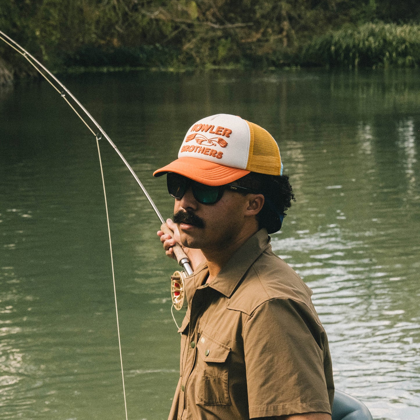 A man wearing sunglasses and a vintage-inspired hat holds a fishing rod while standing near the edge of a calm river, with trees and greenery in the background. - View 2