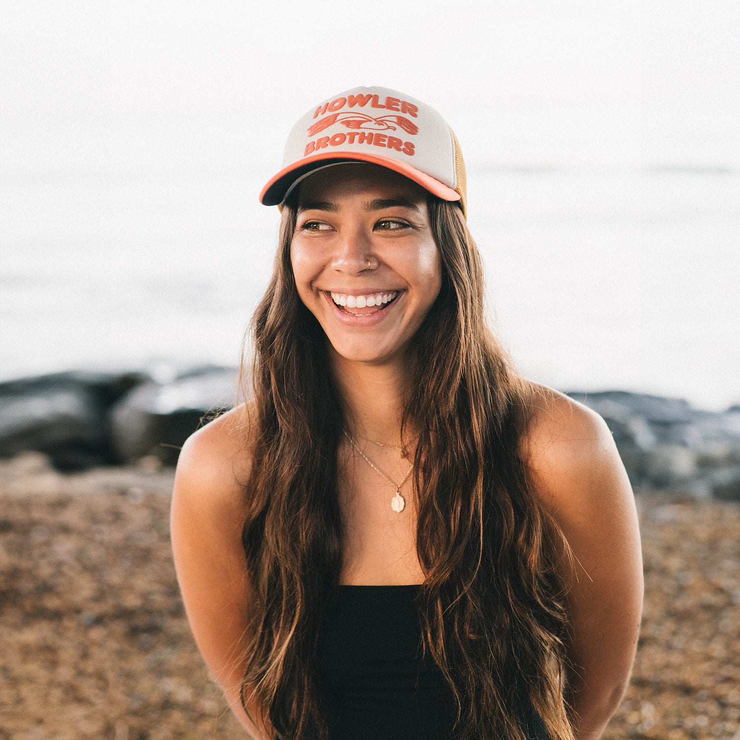 A woman with long brown hair wearing a vintage-inspired Howler Brothers cap and a black strapless top smiles outdoors near a rocky beach, with the ocean blurred in the background. - View 5
