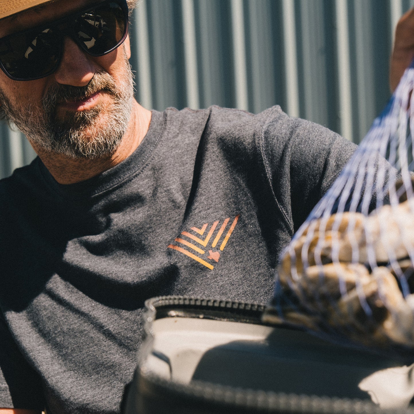 A man wearing sunglasses, a hat, and a JT Van Zandt Collection Howler Select T-Shirt with a geometric design holds a mesh bag filled with what appears to be potatoes outdoors near a corrugated metal wall. - View 2