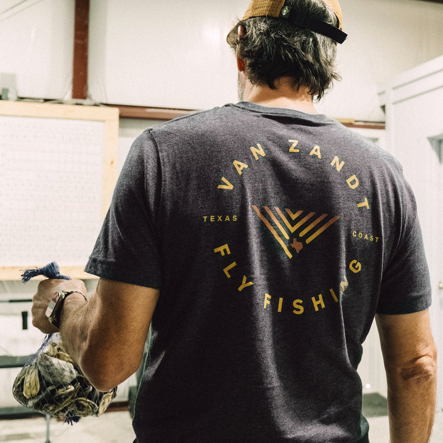 A man in a cap and a Howler Select T-Shirt from the JT Van Zandt Collection stands indoors, holding a drawstring bag in his right hand, facing away from the camera. - View 5