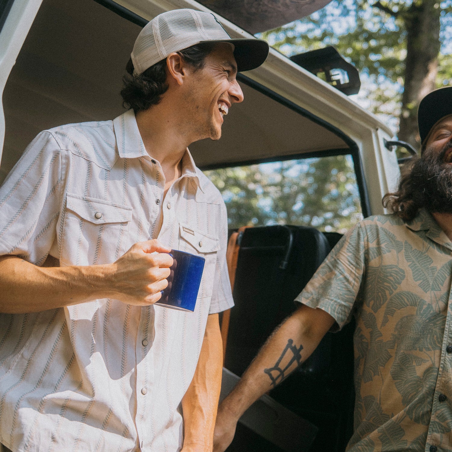 Two men in Howler Brothers vintage inspired shirts and caps laugh together by an open van door in a wooded area. One holds a blue mug, while the other shows a tattoo on his forearm as sunlight filters through the trees. - View 2